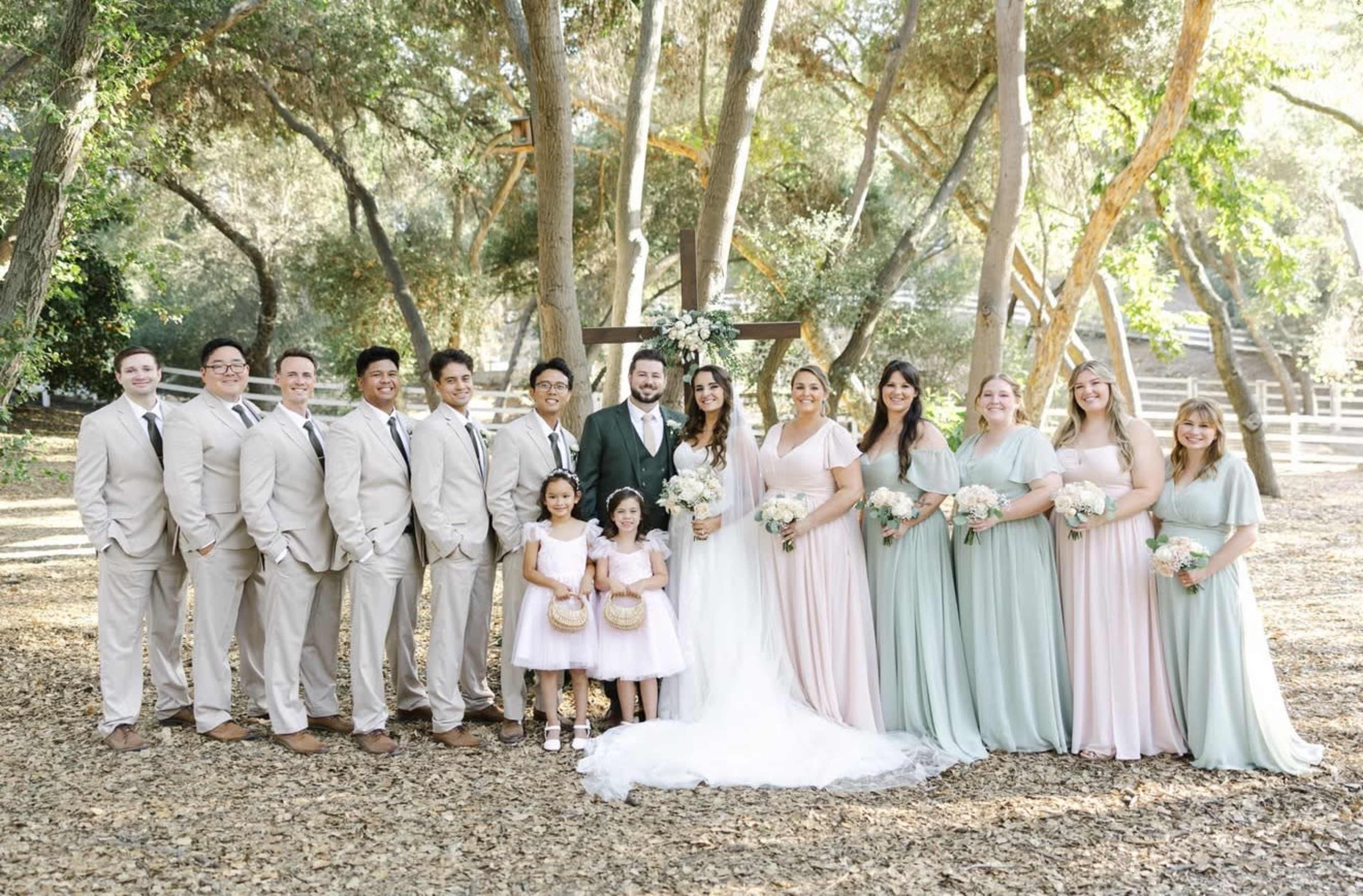 A bridal party stands together in a wooded area, featuring the bride and groom at the center, surrounded by groomsmen, bridesmaids, and two young girls.