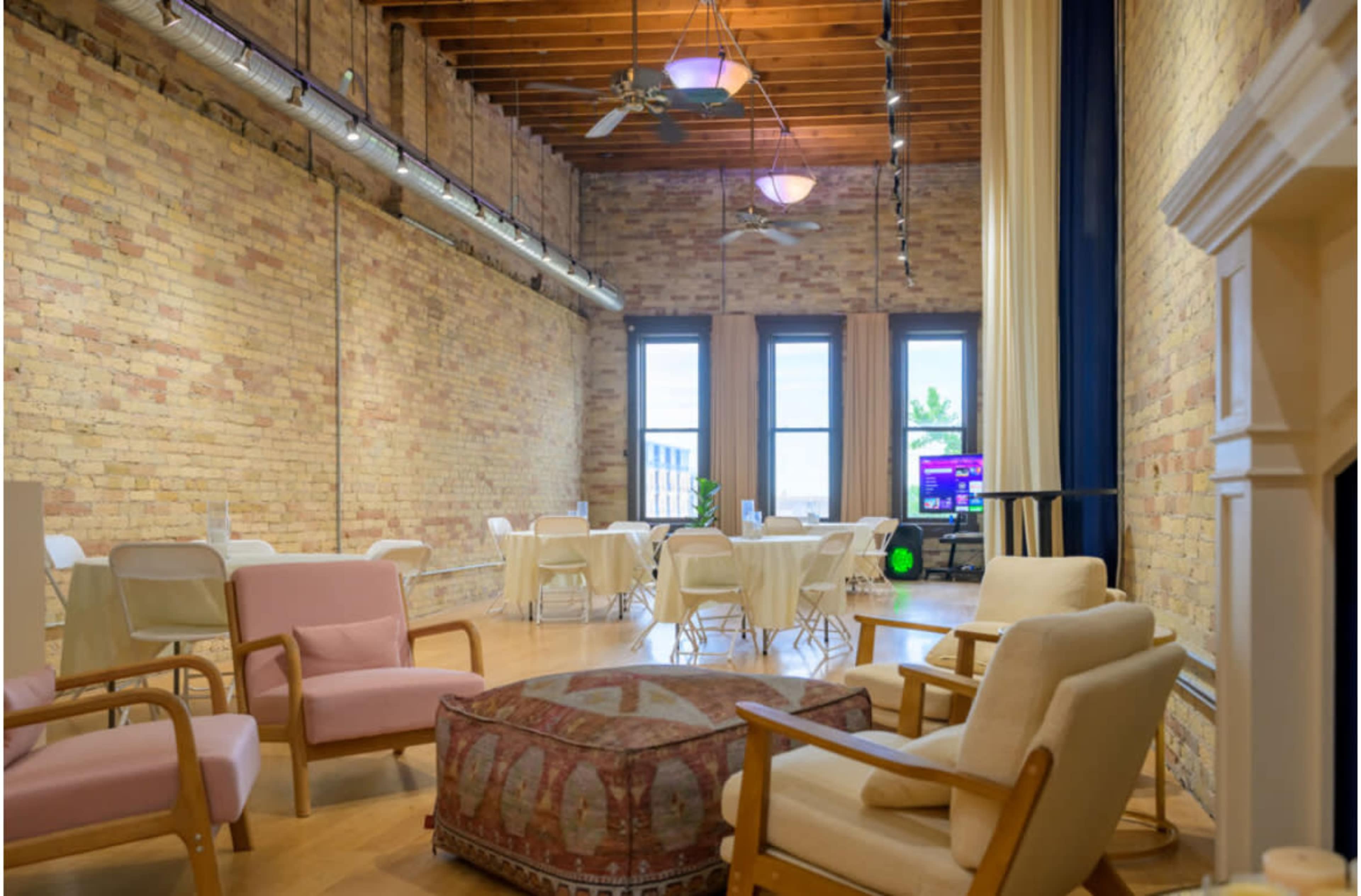 The image shows a spacious room with a high ceiling, featuring exposed brick walls, wooden beams, and several seating arrangements including chairs and tables.
