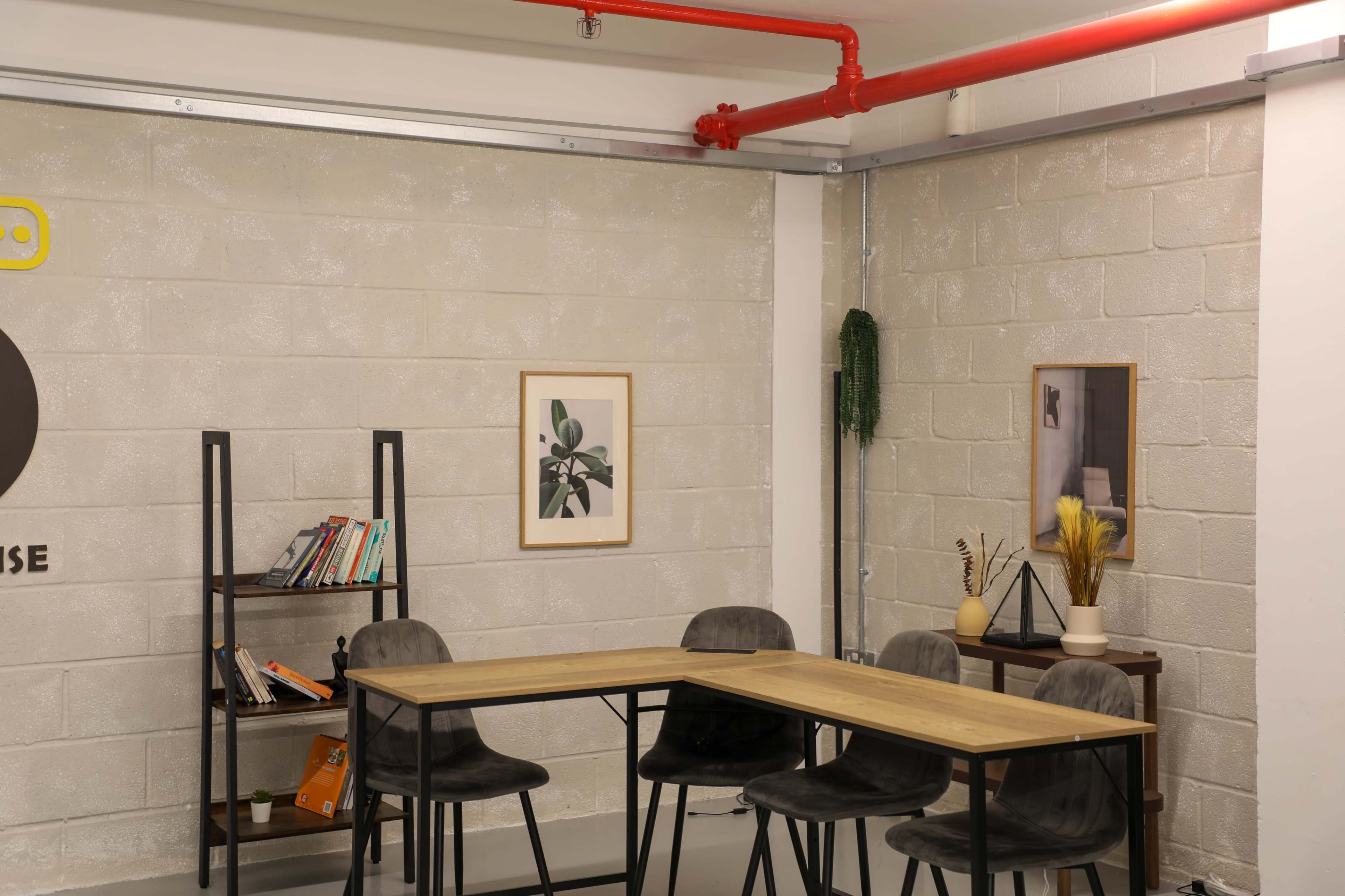 The image shows a minimalist workspace with a wooden table surrounded by four gray chairs, a bookshelf with books, and two framed artworks on the walls.