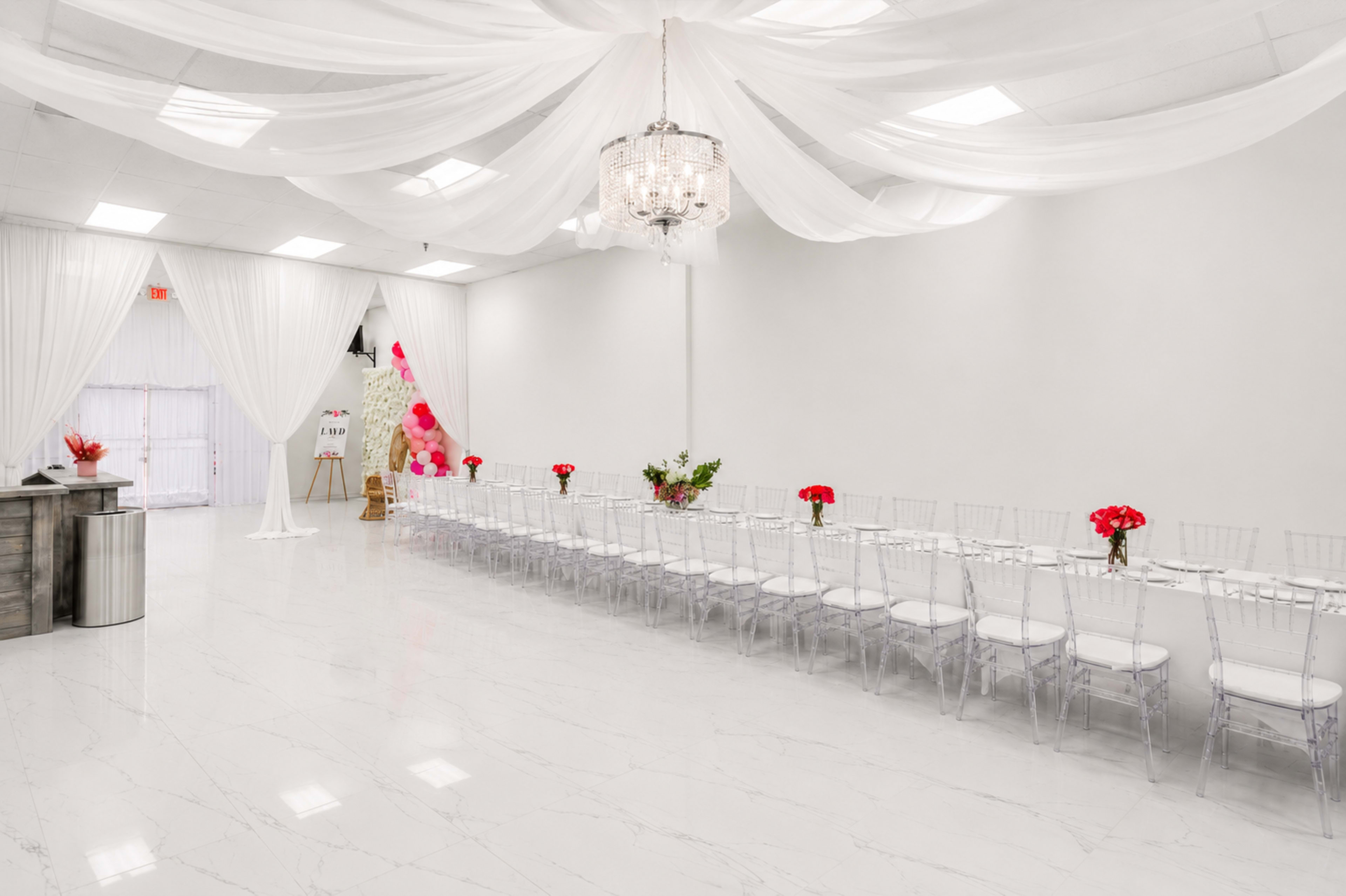 The image shows a spacious, elegantly decorated event room with a long dining table lined with clear chairs and centerpieces of red flowers, beneath a draped ceiling with a chandelier.