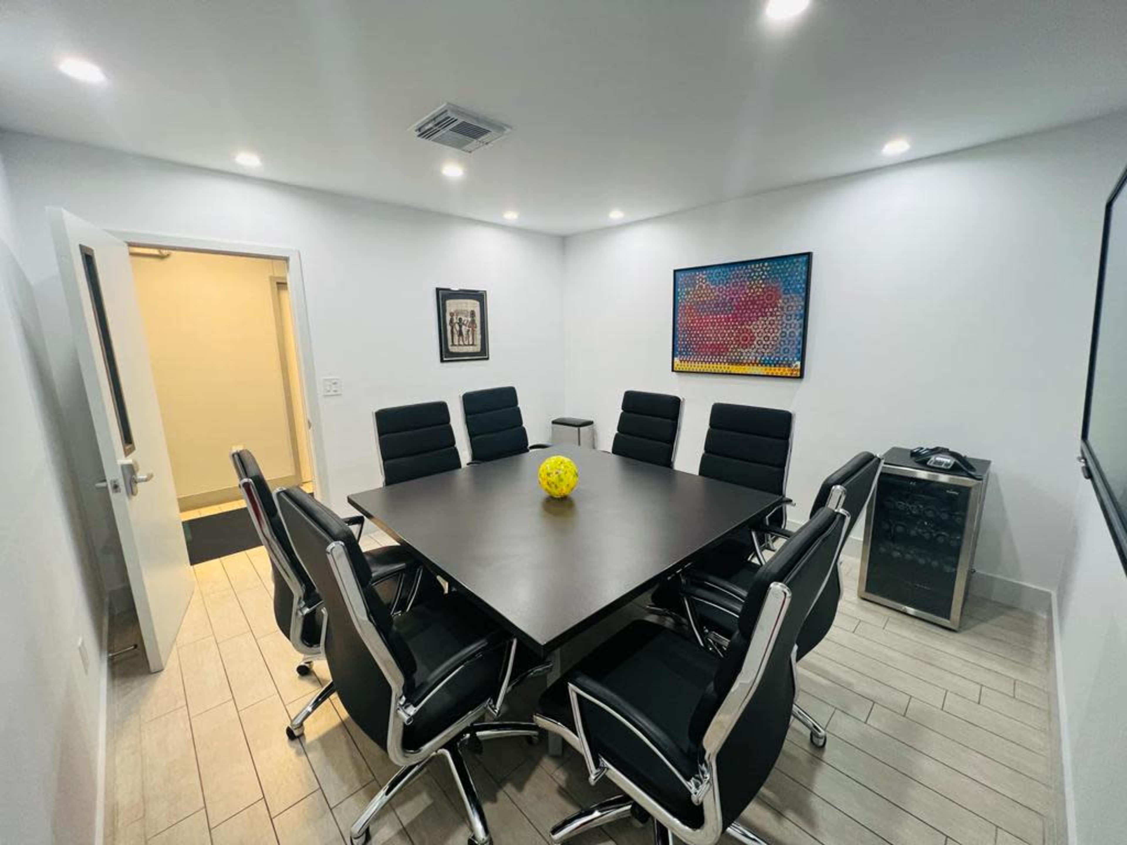 A conference room features a large rectangular table surrounded by black chairs, with a yellow decorative ball at the center and a wine fridge in the corner.