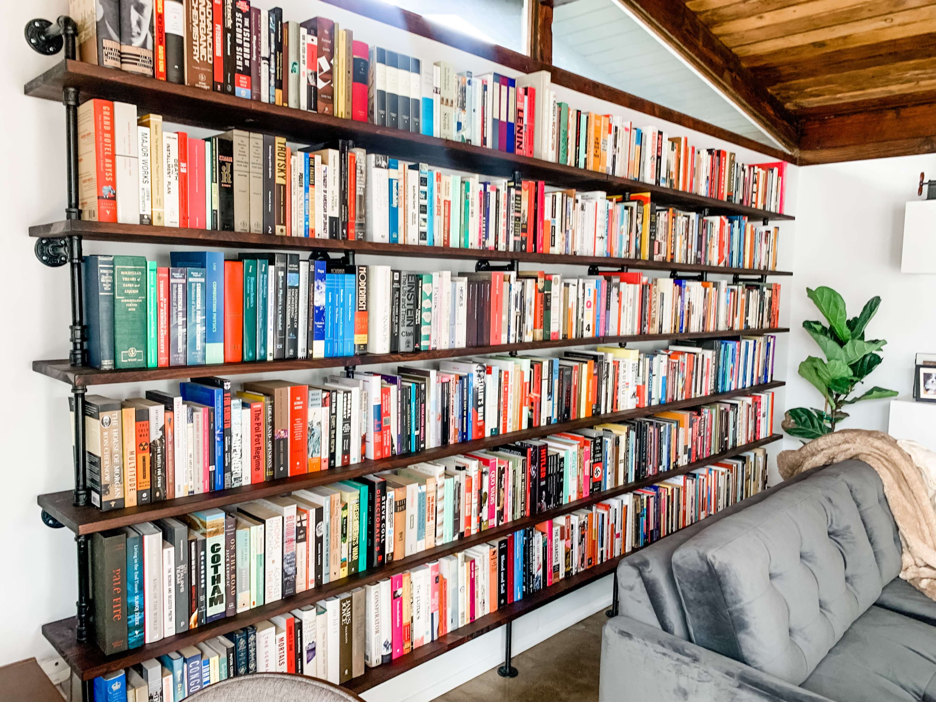 A wooden bookshelf filled with a wide variety of books, positioned next to a gray couch in a room with wooden beams.