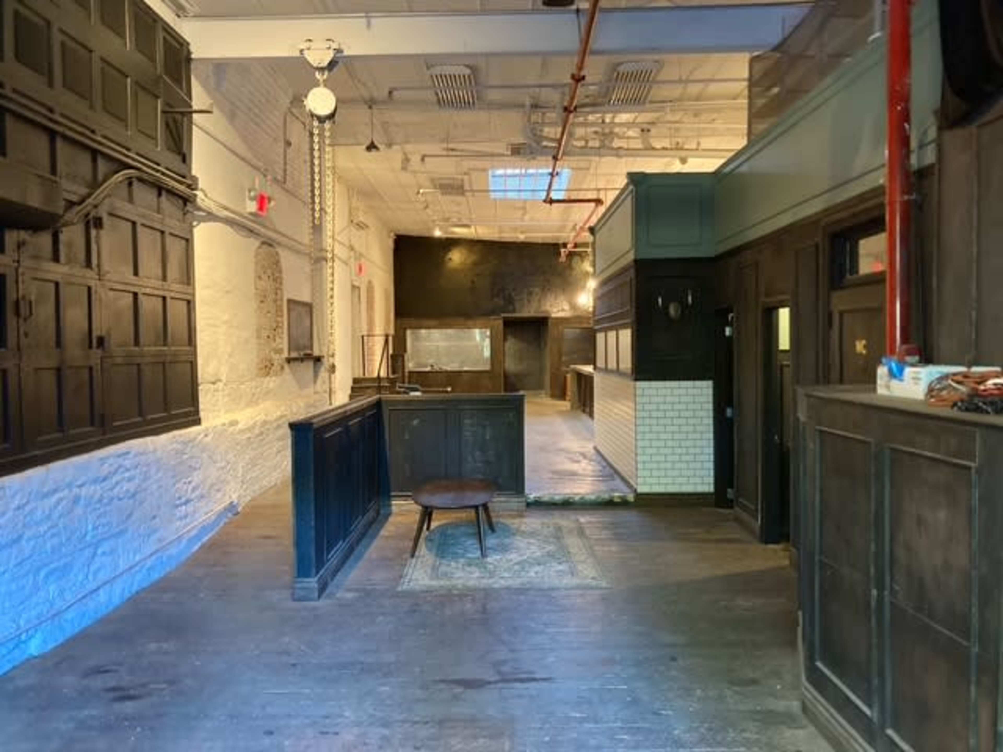 The image shows an interior space with wooden flooring, a central table, and a combination of dark wood and light-colored walls, featuring a large window on the ceiling.