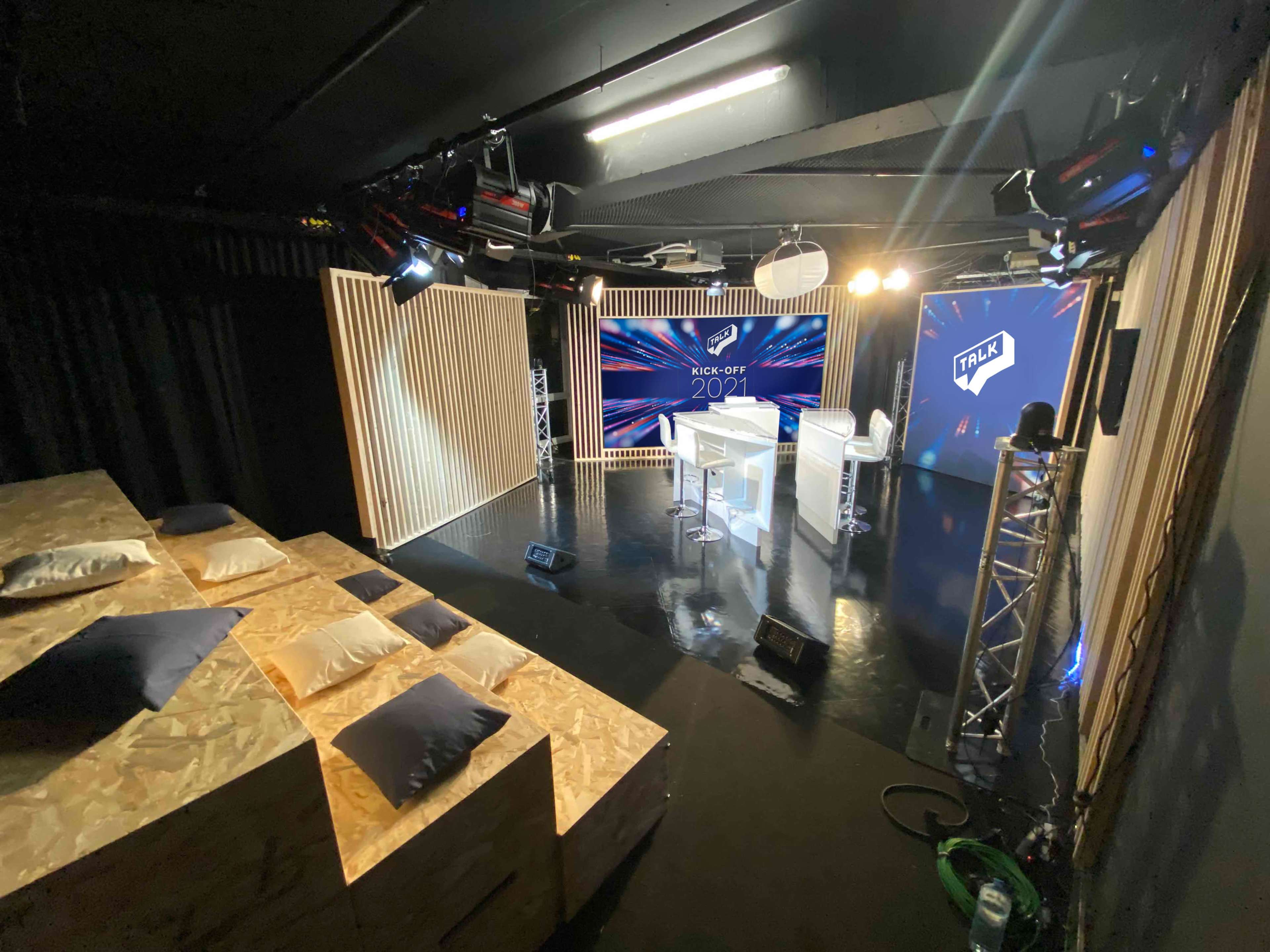 A modern studio setup with a central table and seating area, illuminated by stage lights, and featuring branded screens in the background.