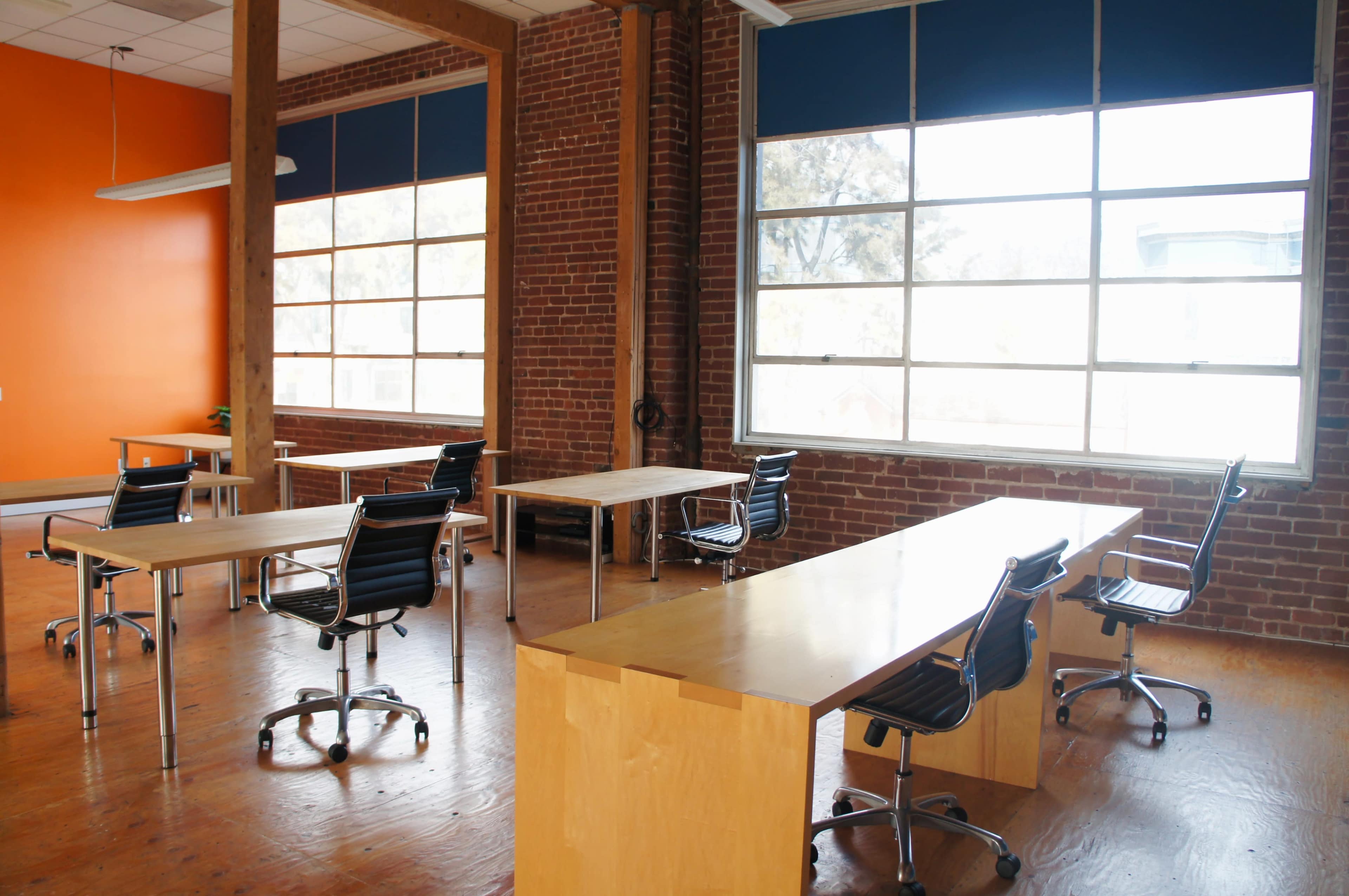 A bright, spacious room features several wooden tables and rolling chairs, with large windows allowing natural light to illuminate the brick walls.