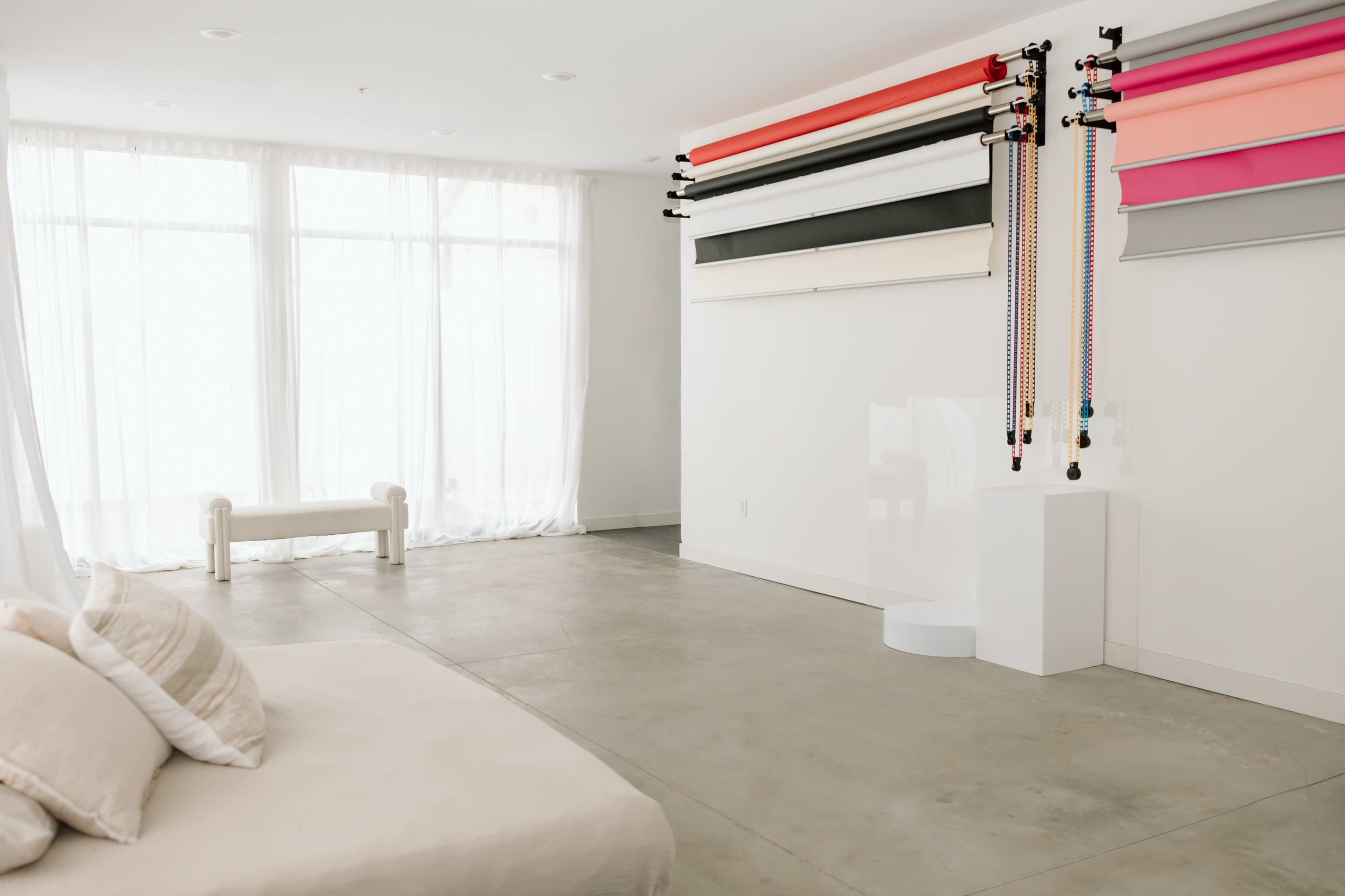 The image shows a modern, minimalist studio space with a large window, concrete flooring, and various colored backdrop rolls mounted on the wall.