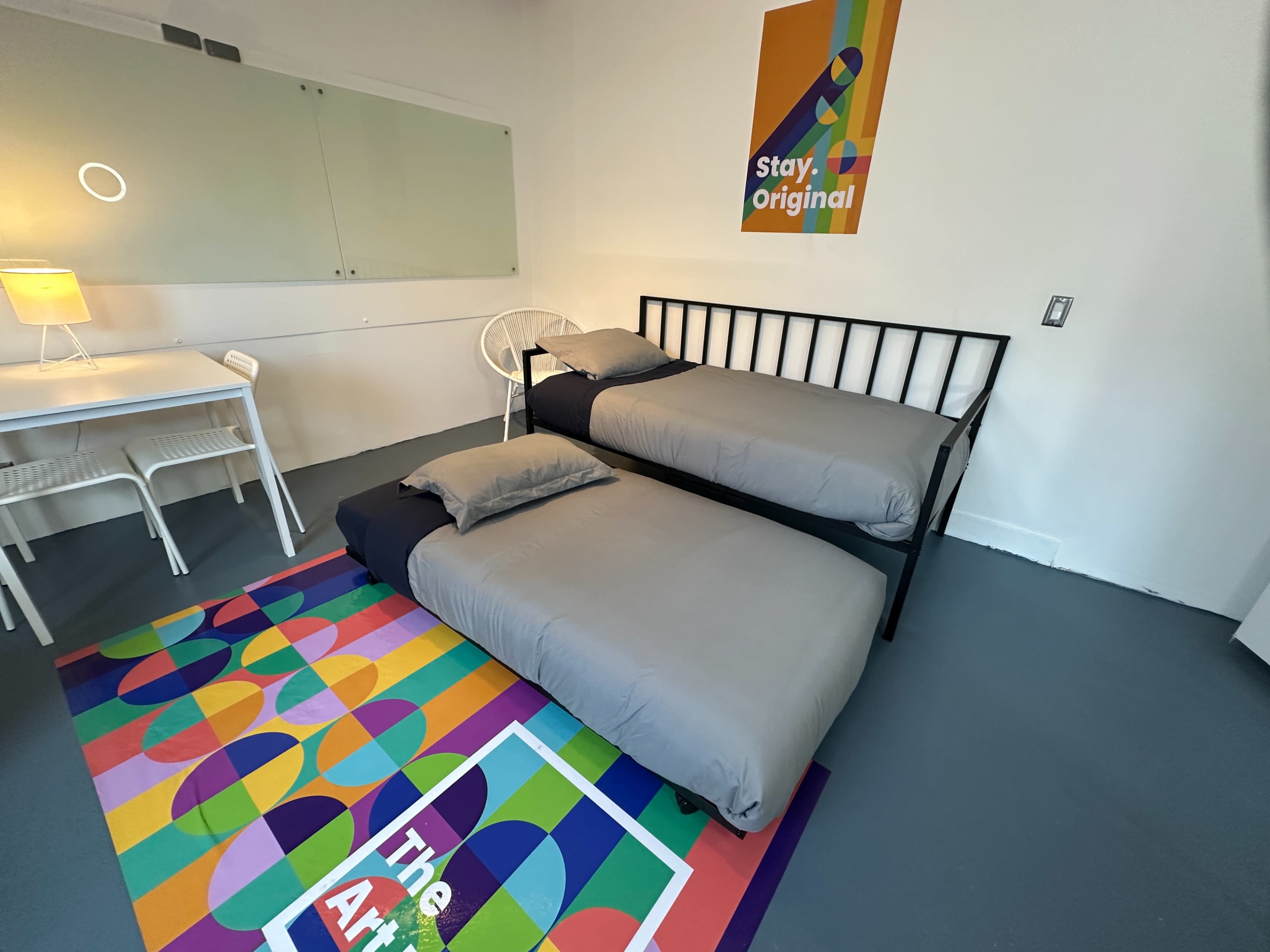 The image shows a minimalist bedroom with two beds, a small table with a lamp, and a colorful rug on the floor.