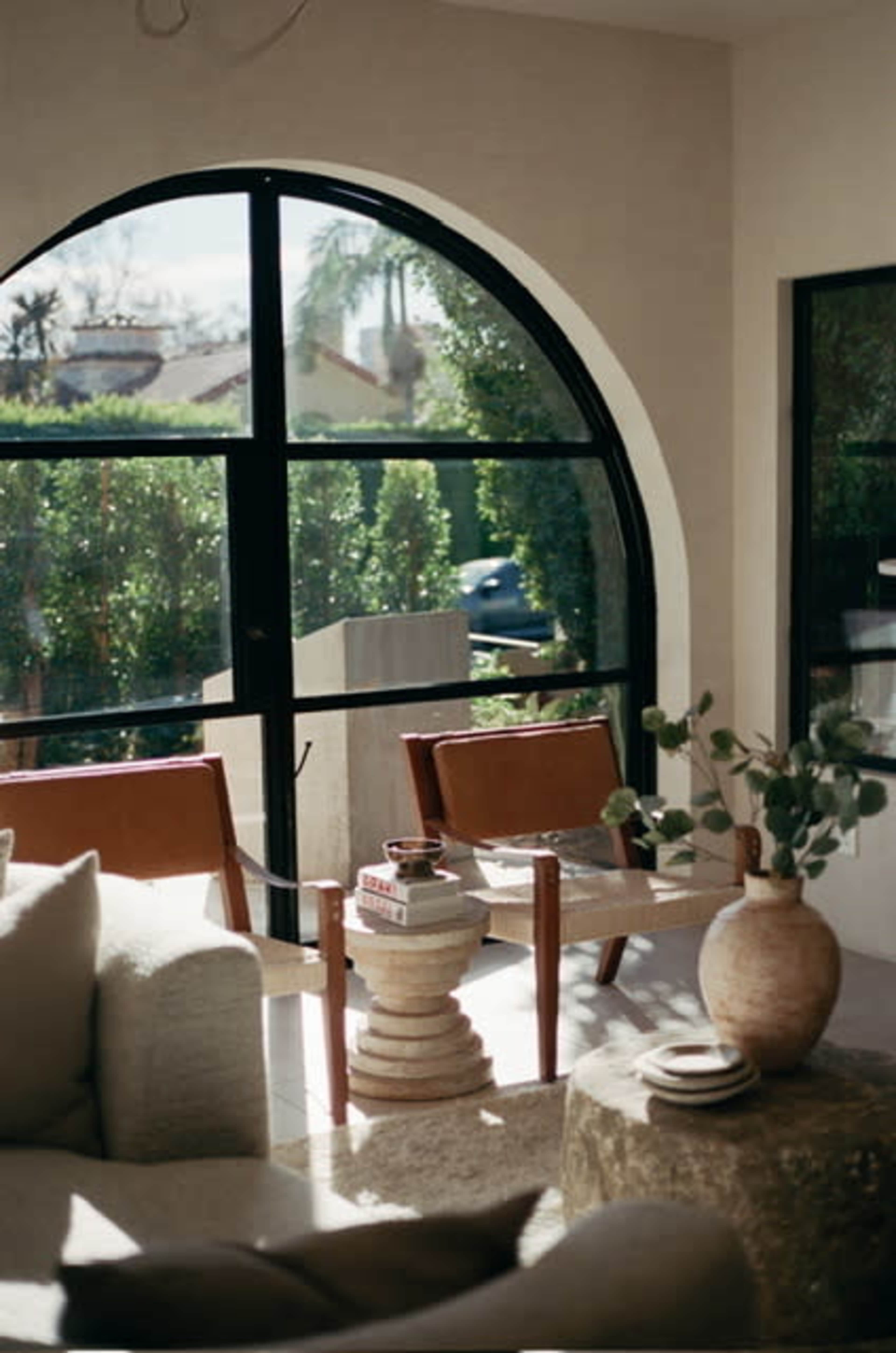 The image shows a modern interior space featuring two brown chairs, a stack of decorative items, and a large window that offers a view of a landscaped garden.