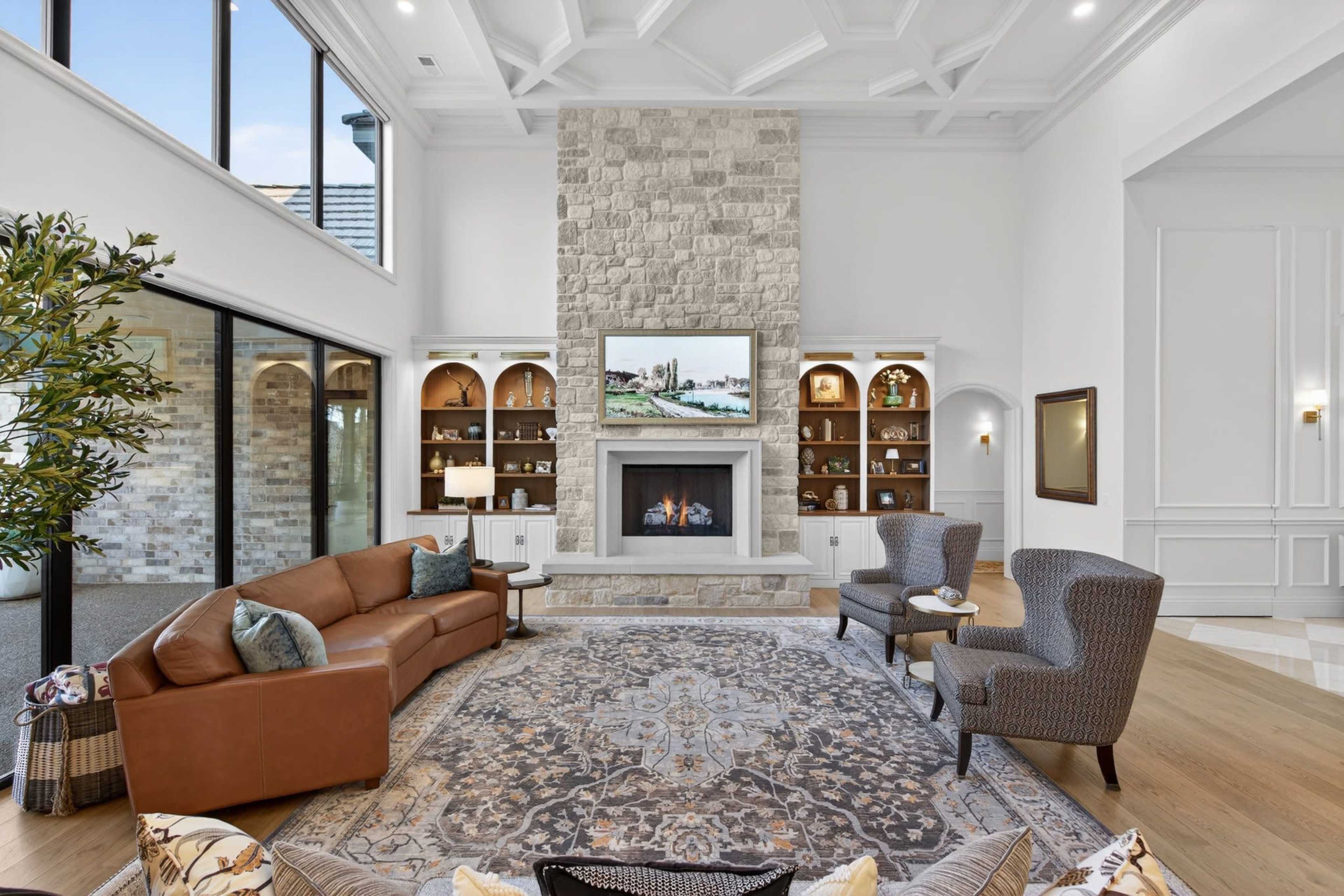 The image shows a spacious living room with a stone fireplace, built-in shelves, and large windows allowing plenty of natural light.