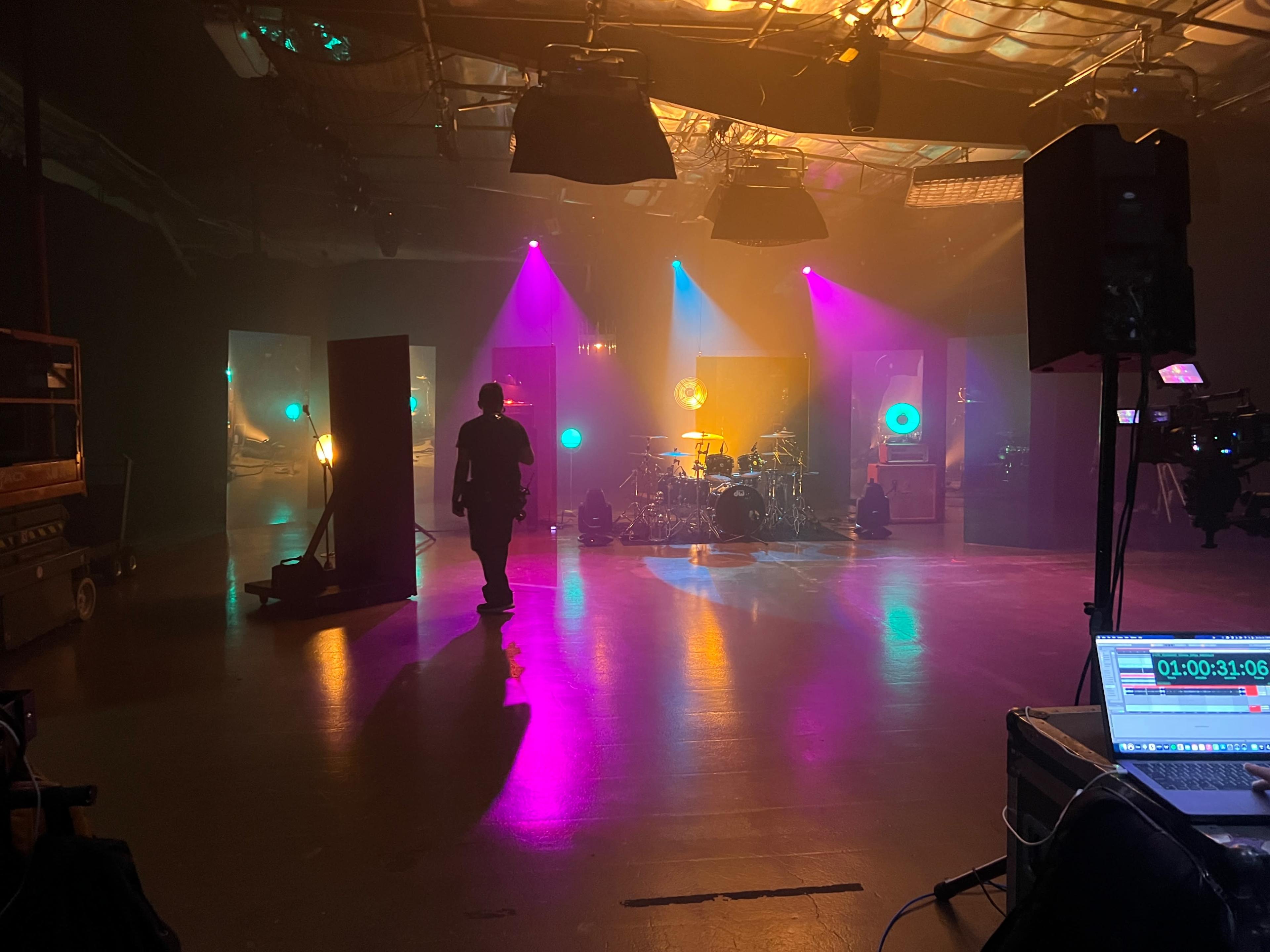 A person walks through a dimly lit studio set with colorful stage lights illuminating the space and a drum set in the background.