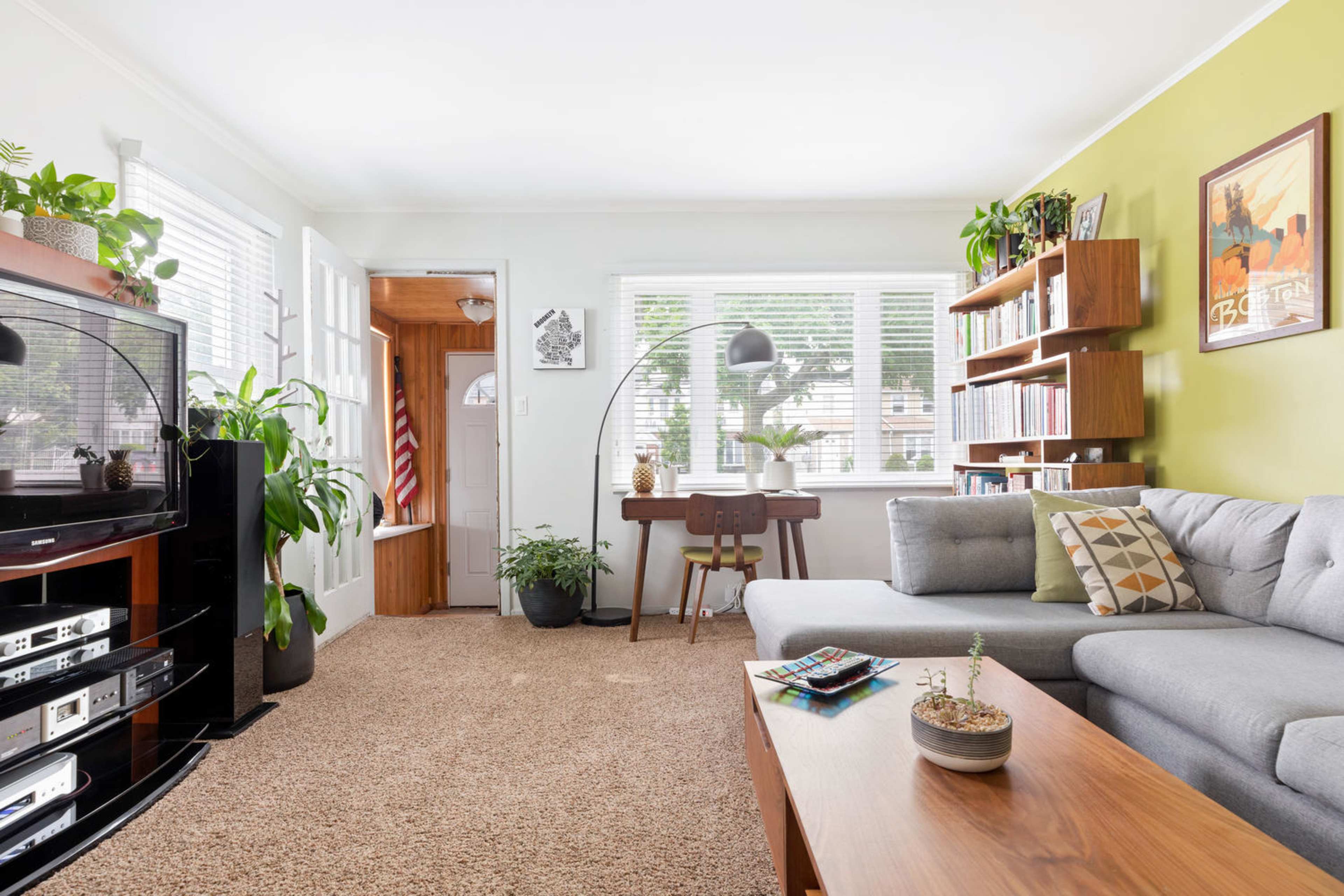 A bright living room featuring a gray sofa, a wooden coffee table, and a large television, with plants and bookshelves adding decorative elements.