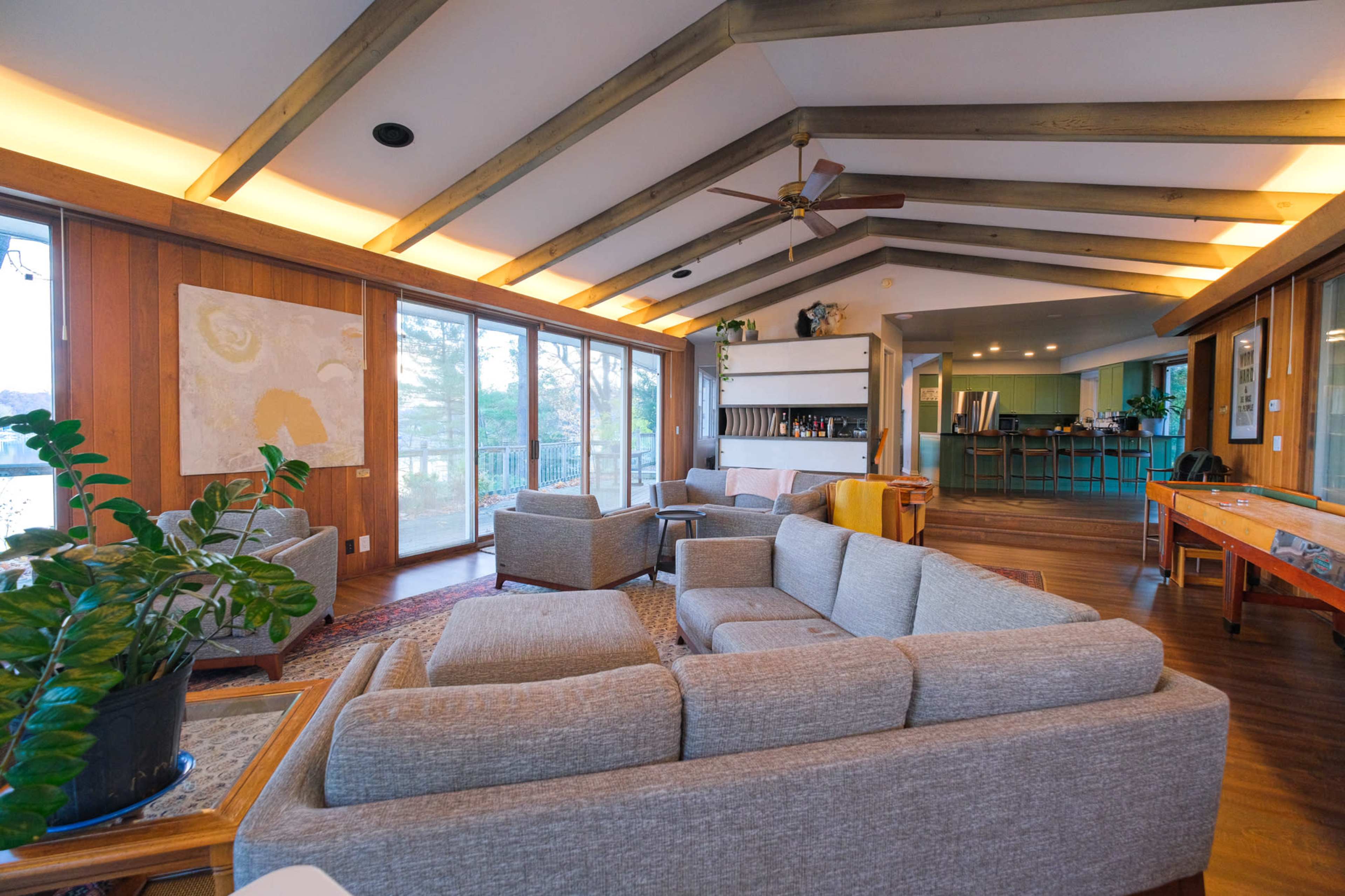 A modern living room with a light gray sectional sofa, wooden beams on the ceiling, and large windows overlooking a natural view.