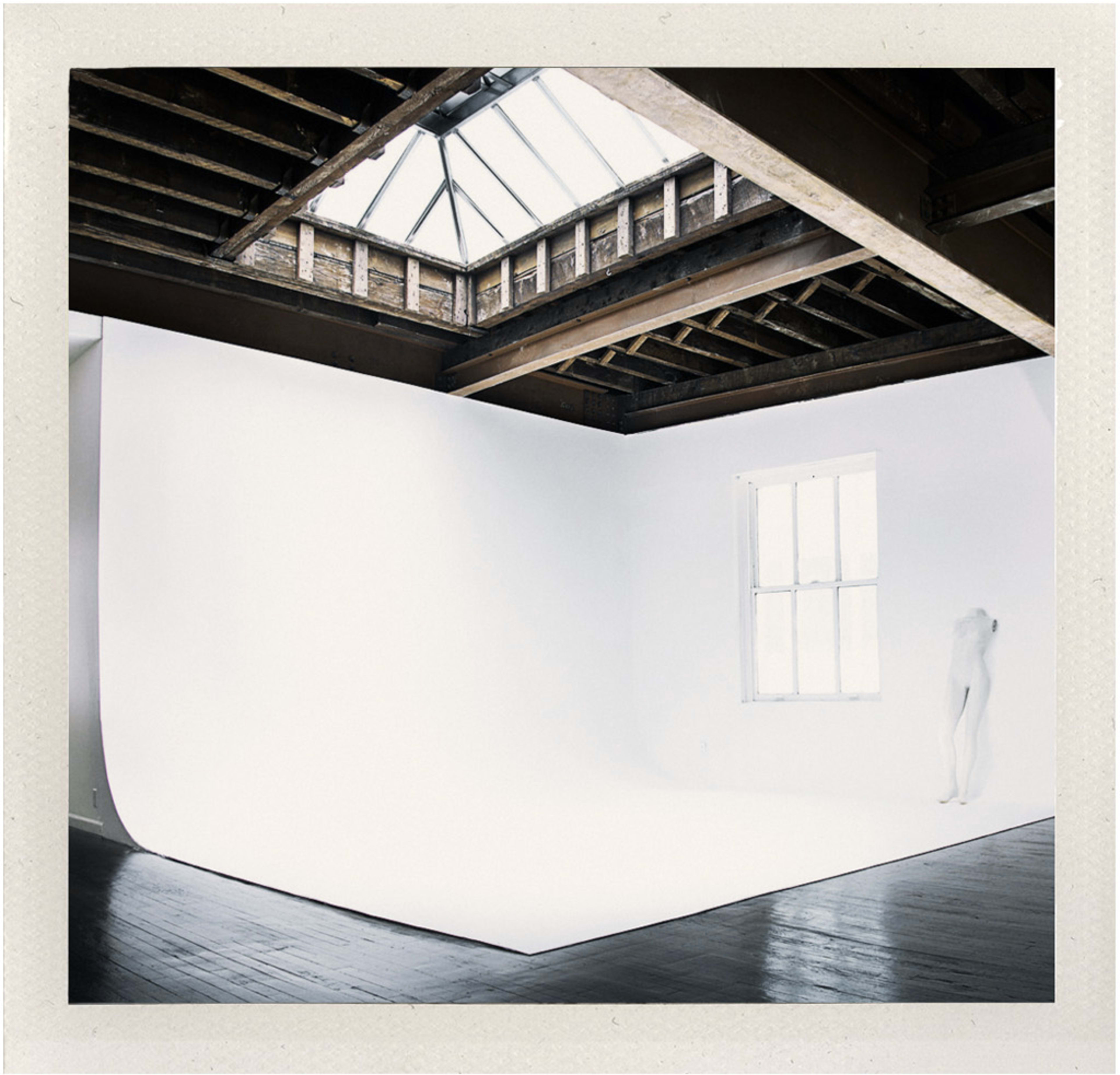 A bright, minimalistic studio space features a white backdrop, wooden beams, and a skylight illuminating the interior.