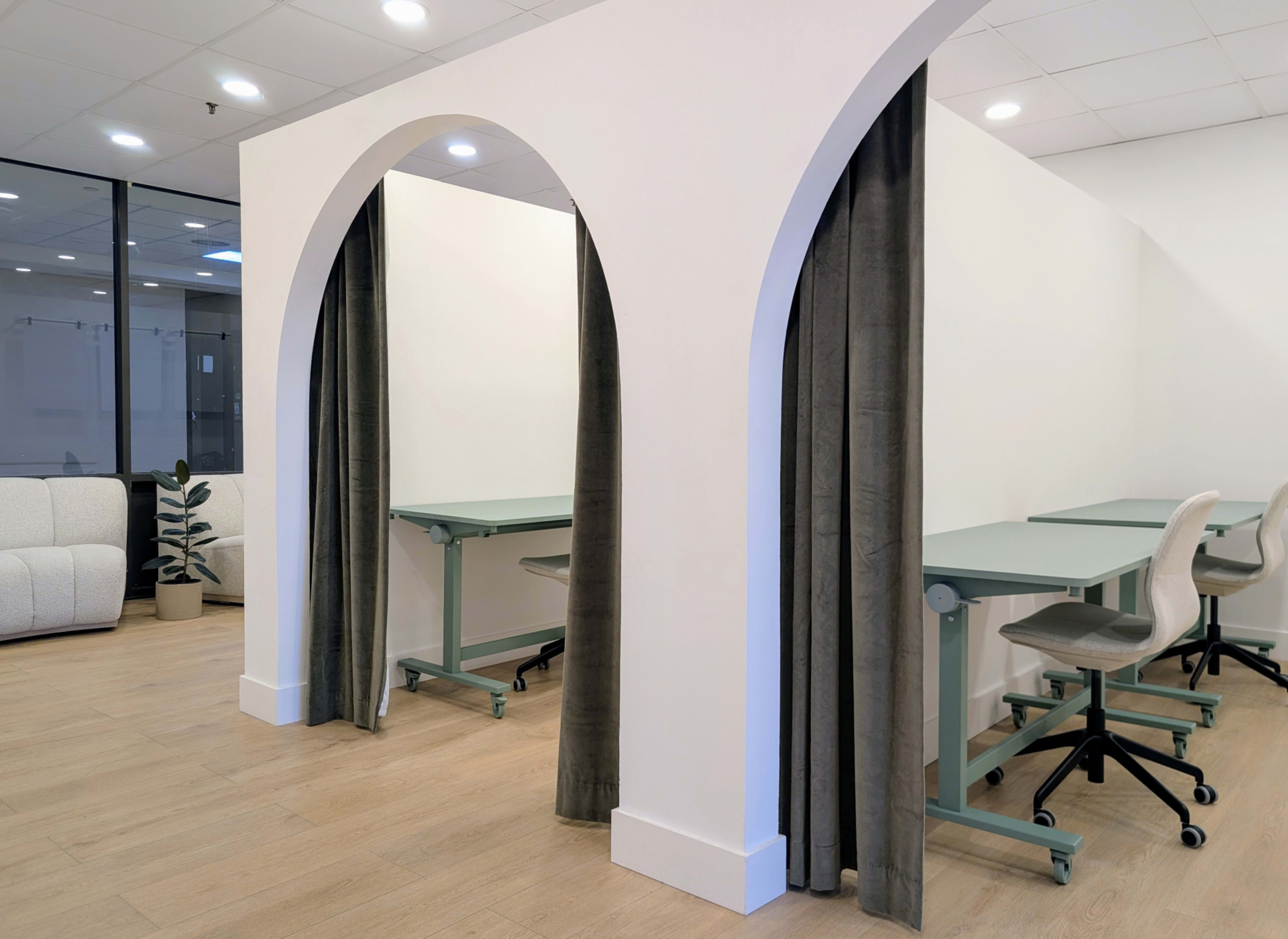 The image shows a modern workspace featuring arched openings with curtains leading to individual workstations, each equipped with a desk and a chair.