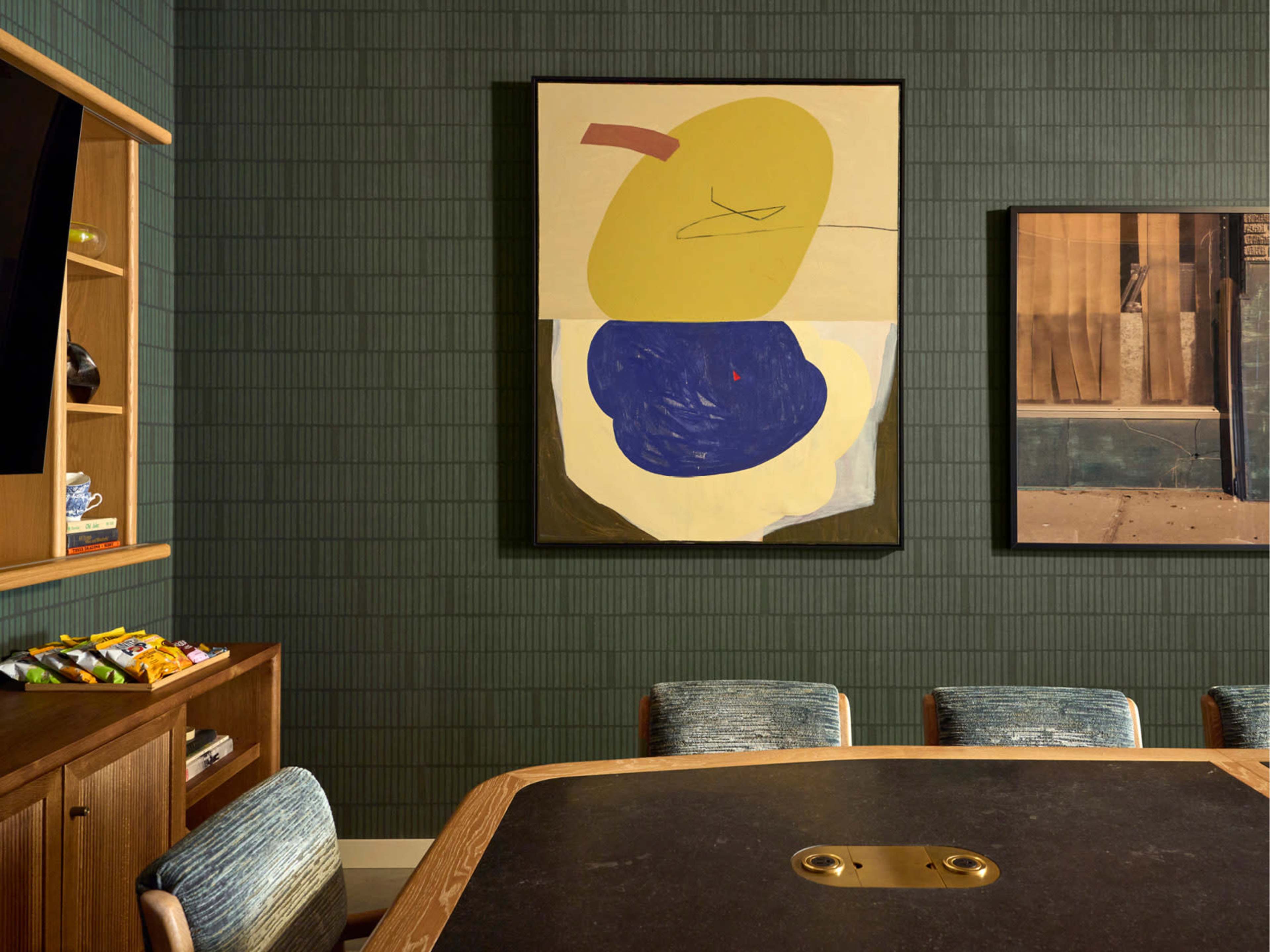 The image shows a modern conference room with a dark green textured wall, a round table, and two abstract artworks displayed on the walls.