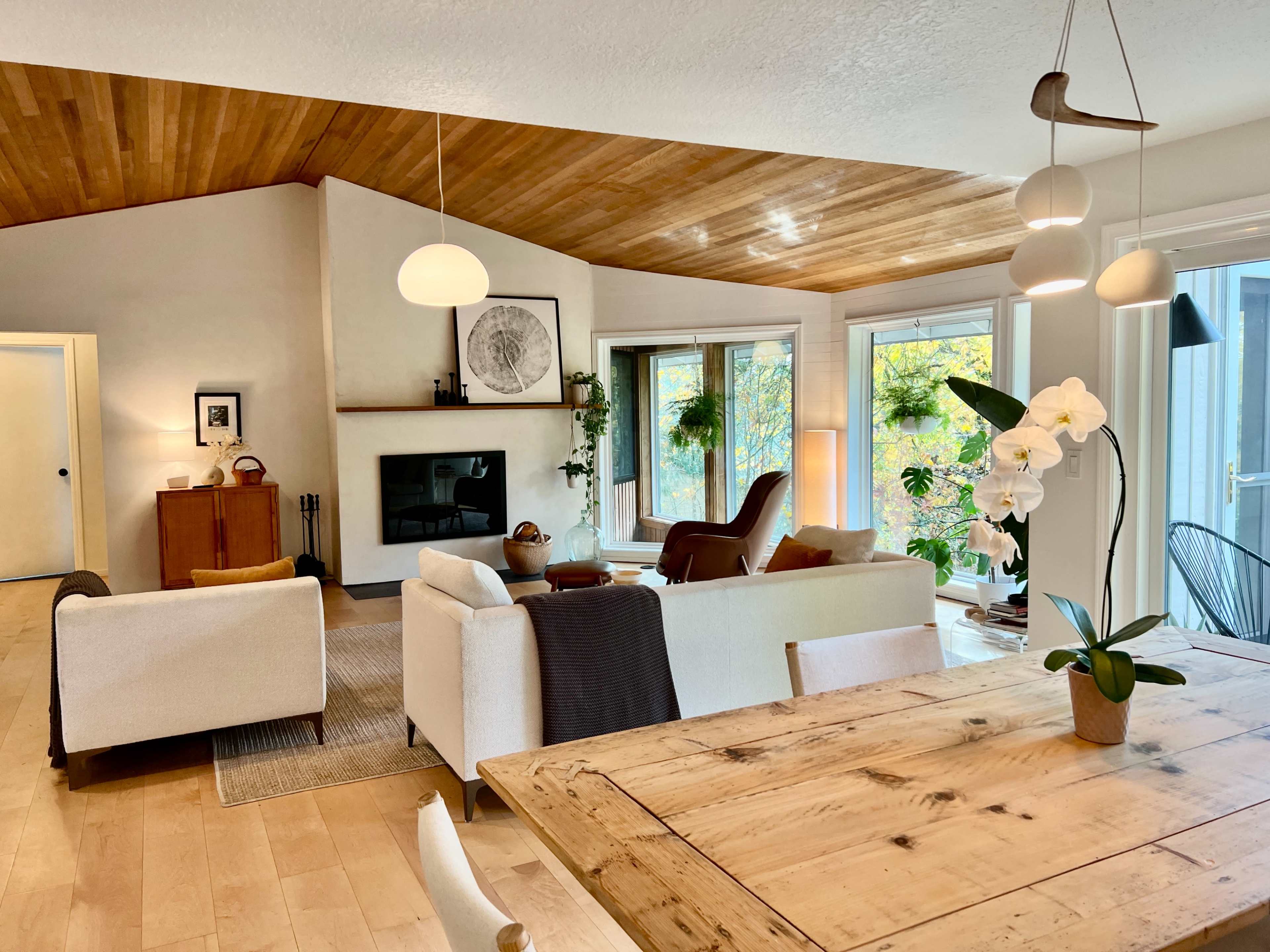 A modern living room with a light wooden ceiling, minimalist furniture, and large windows allowing natural light to filter in.
