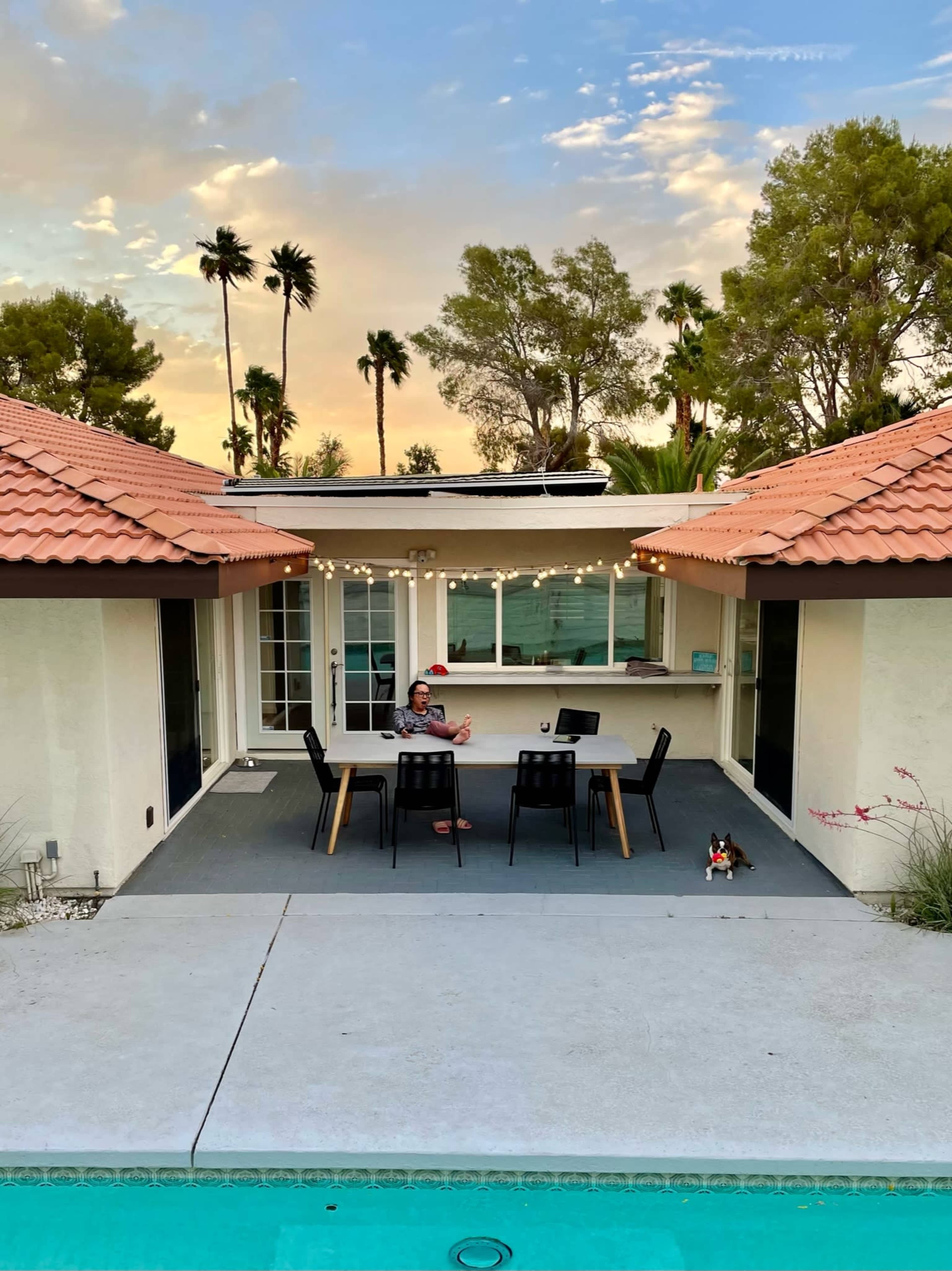 A person sits at a dining table on a patio between two rooftops, with a dog nearby, overlooking a pool and palm trees under a colorful sky.