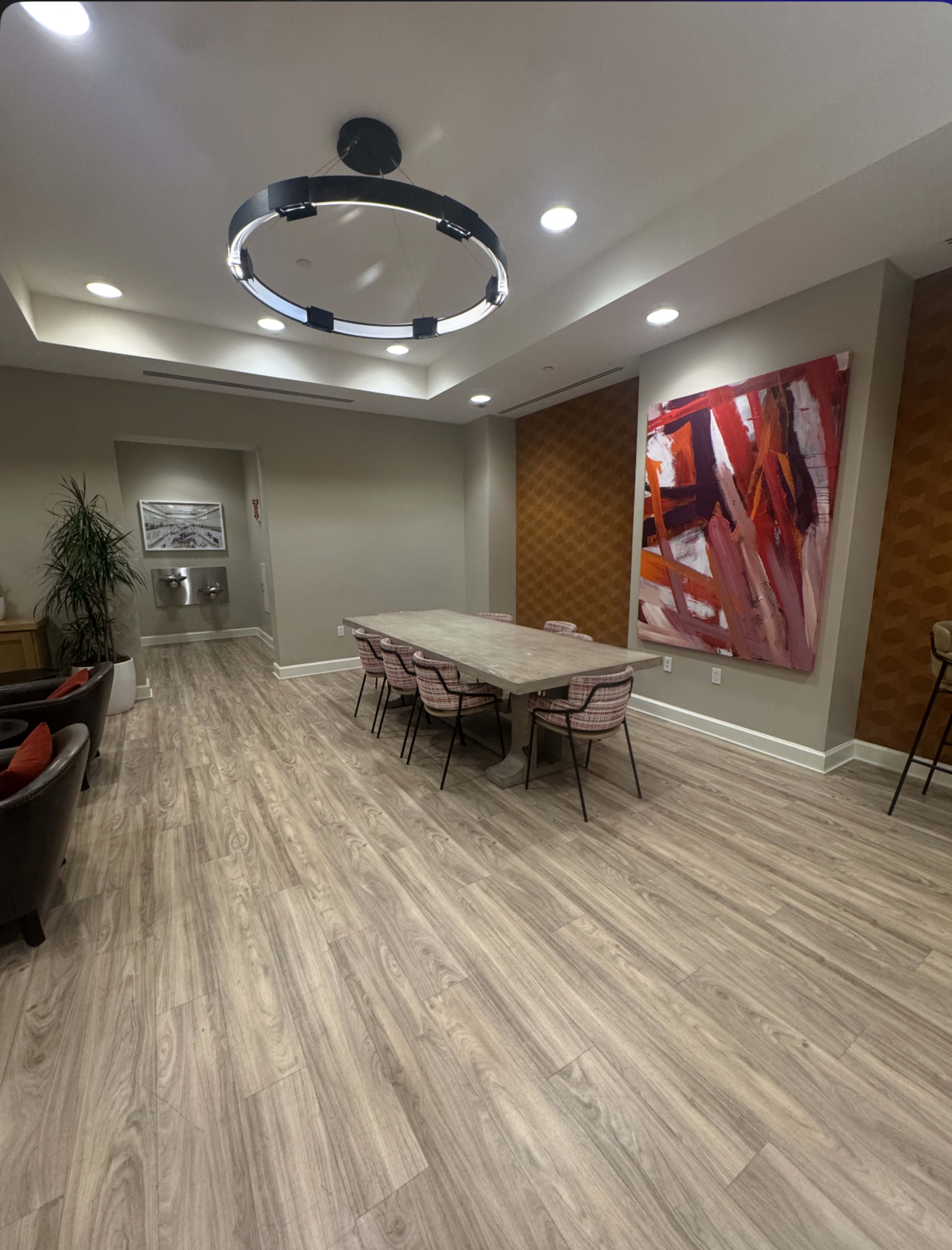 The image shows a modern meeting room featuring a long wooden table surrounded by chairs, with a large colorful abstract painting on one wall.