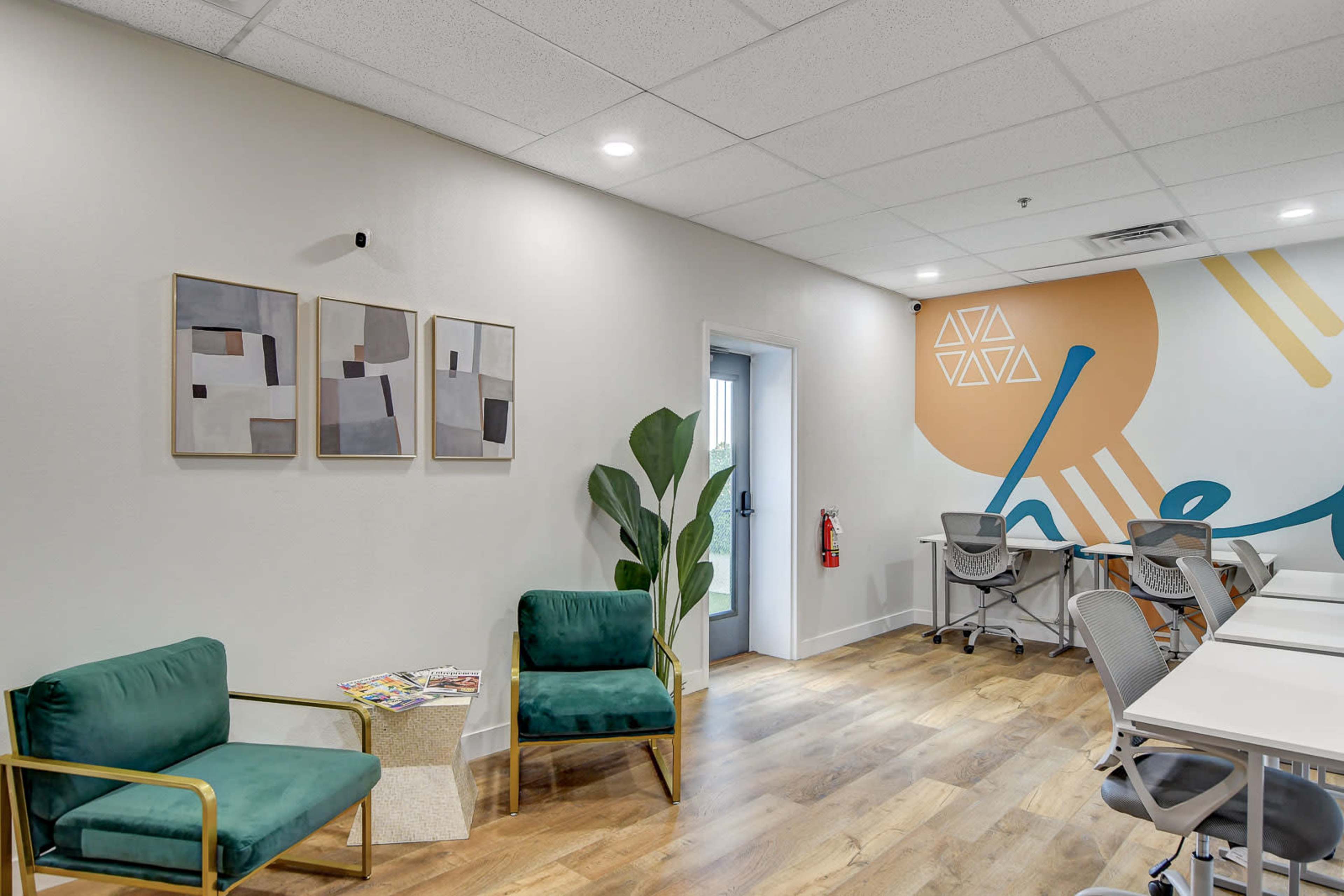 A modern co-working space featuring two green chairs, a small table with magazines, and a colorful accent wall with geometric designs.