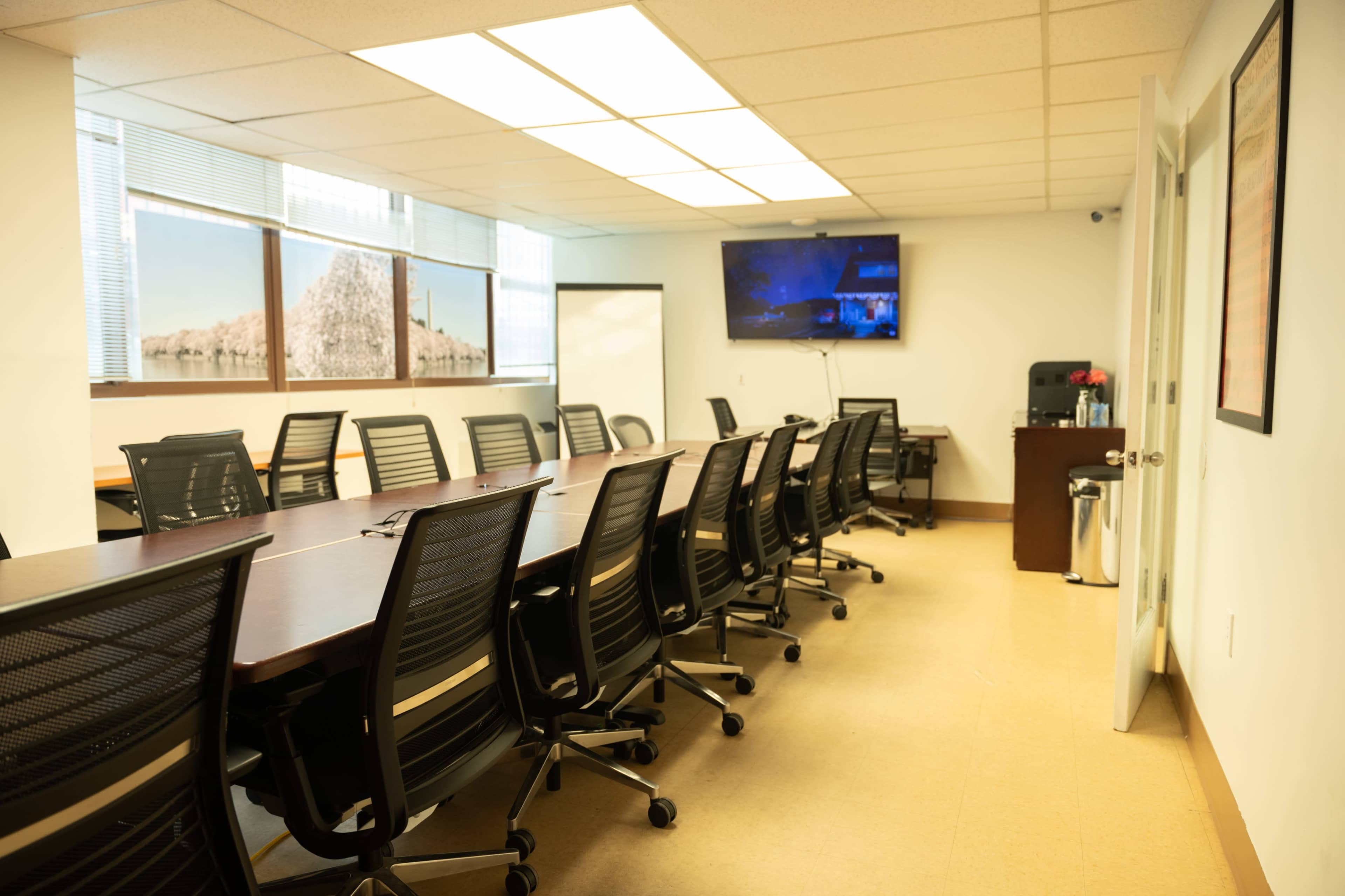 A conference room with a long table and multiple black chairs, featuring a large screen and windows with a view.