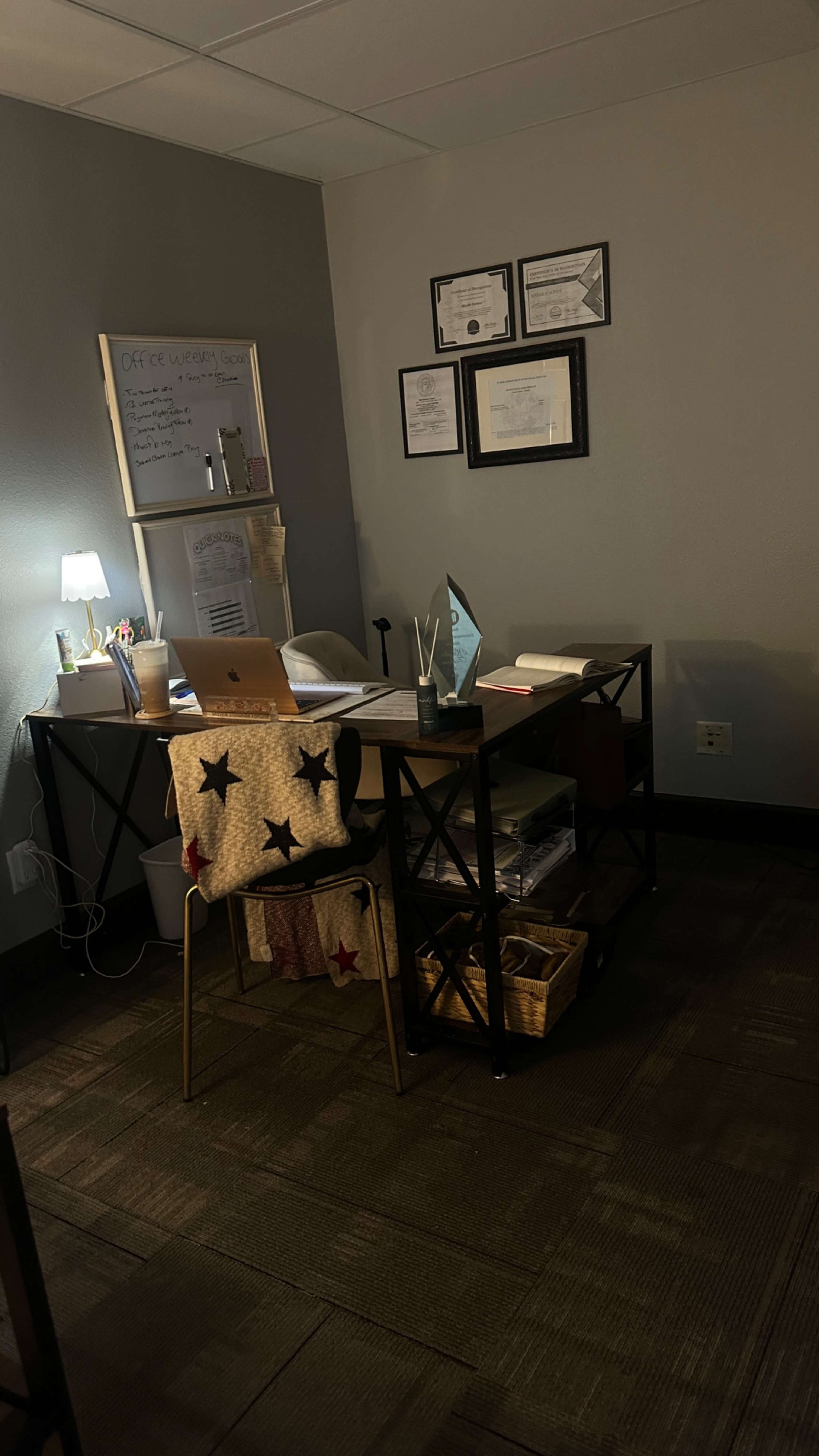 The image shows a small office space with a desk, a laptop, a lamp, and various documents and decorations on the walls.