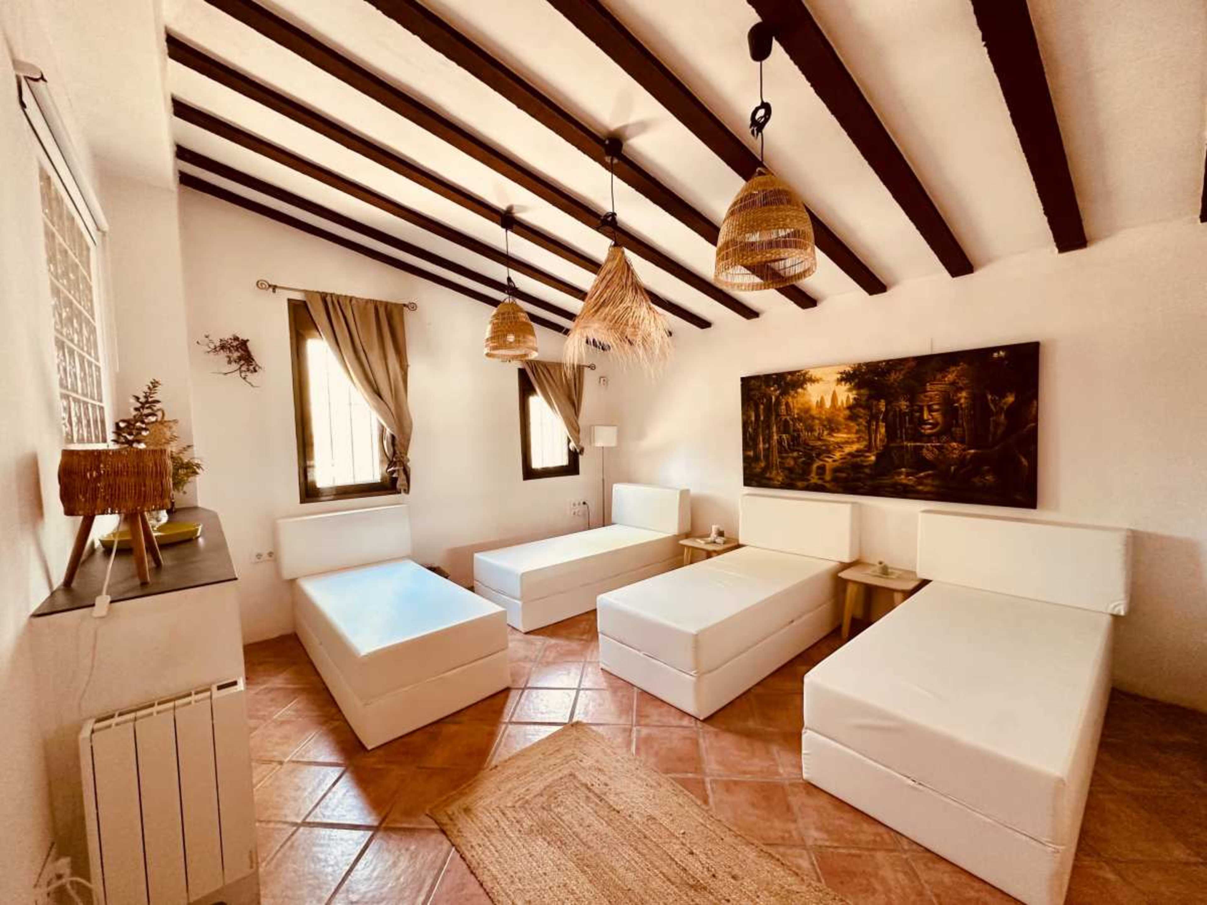 The image shows a room with four white beds arranged around the floor, featuring wooden beams overhead and decorative lighting.