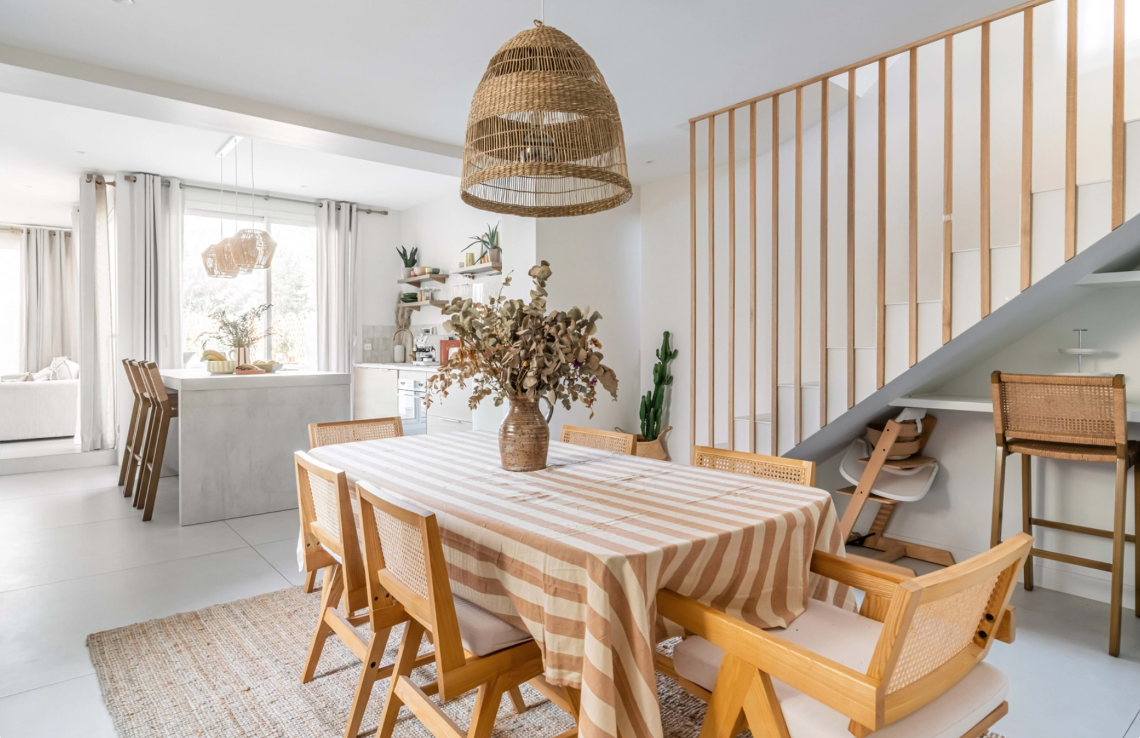 The dining area features a wooden table surrounded by chairs, adorned with a striped tablecloth, with a staircase and kitchen visible in the background.