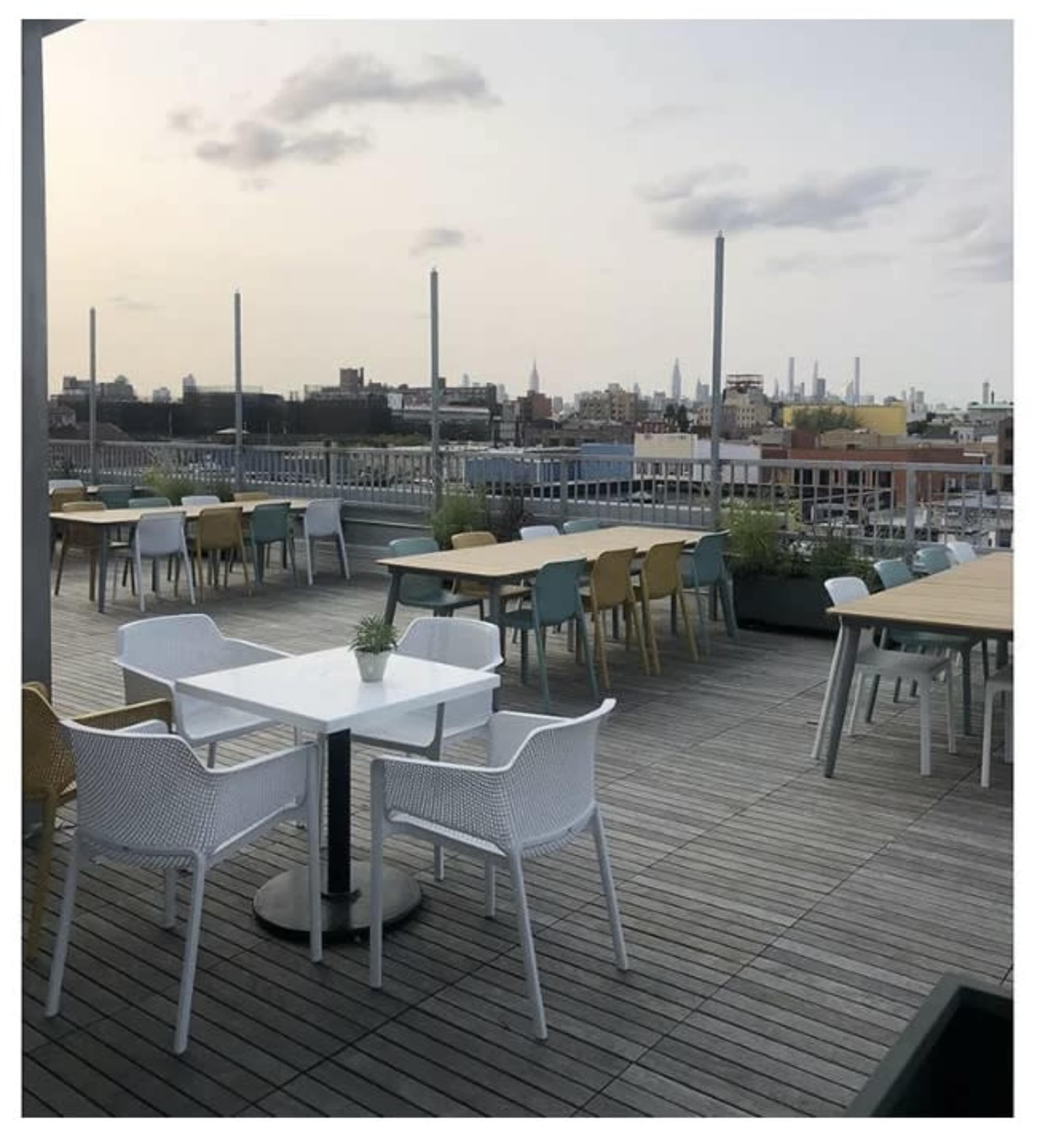A rooftop terrace features multiple tables and chairs with a view of a city skyline at dusk.
