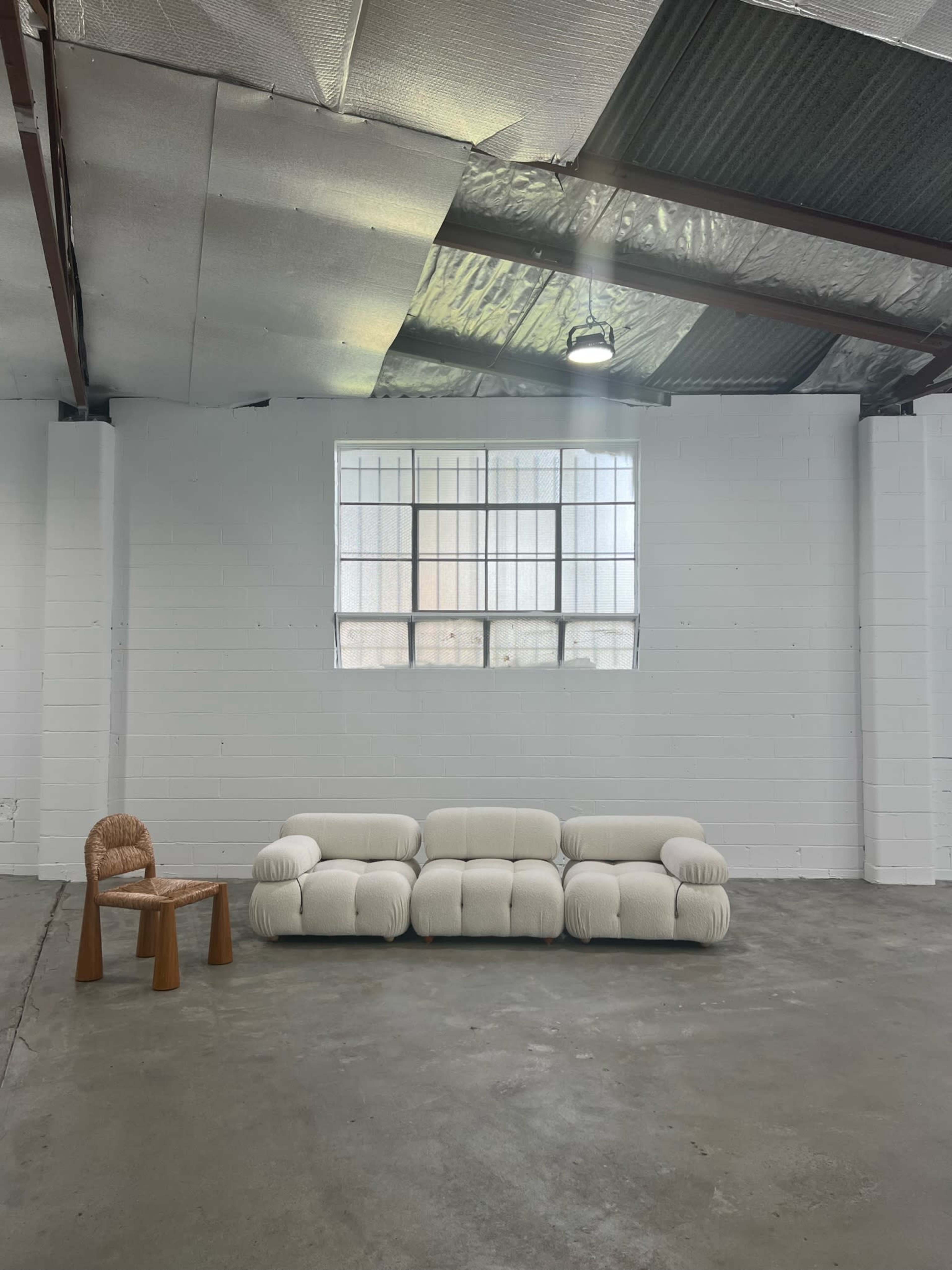 A white, plush sofa is positioned against a bare wall beneath a large window, alongside a wooden chair in an industrial space with a concrete floor.