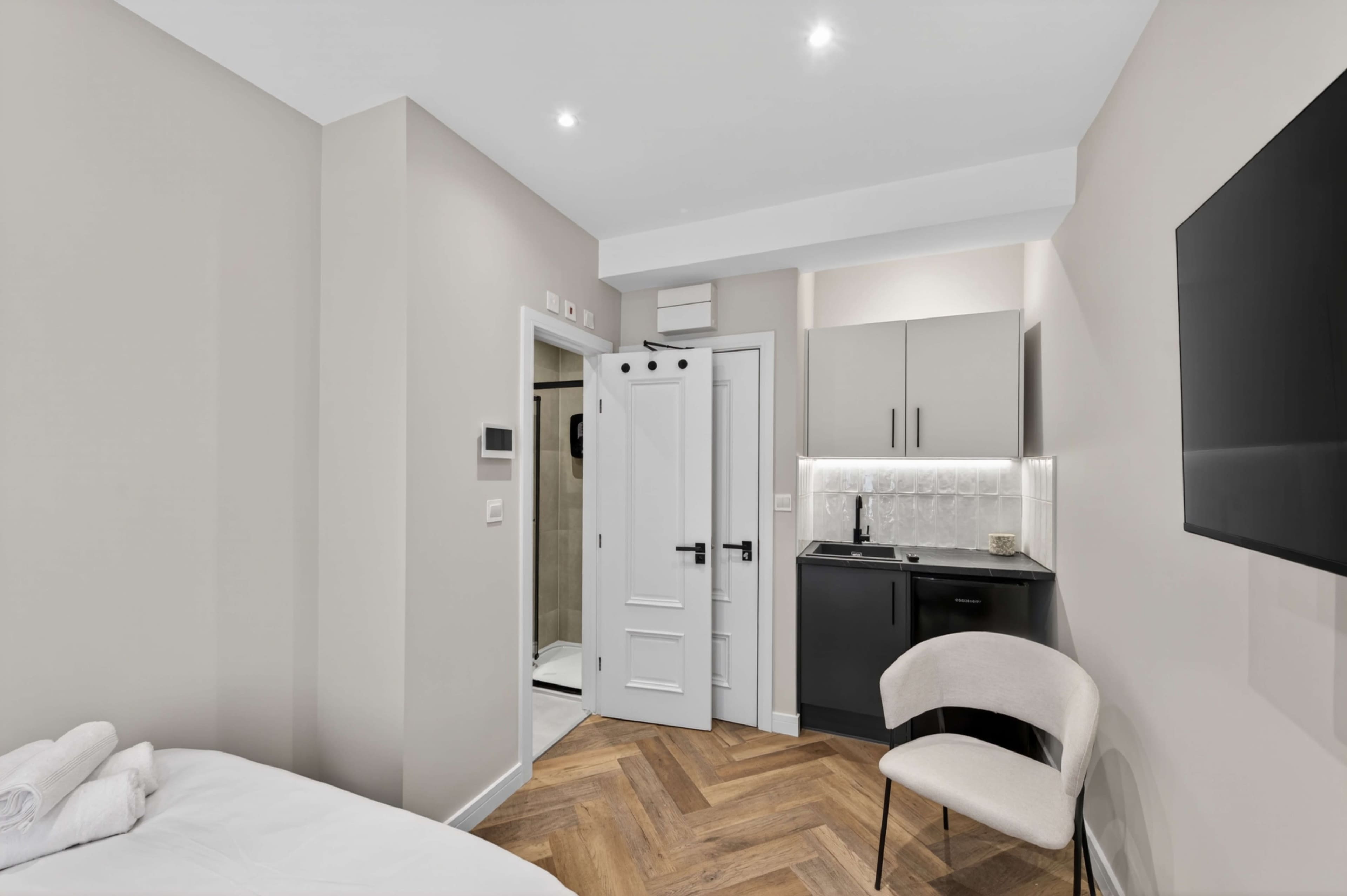 The image shows a modern hotel room featuring a kitchenette, a bathroom door, and a single chair against a light-colored wall.