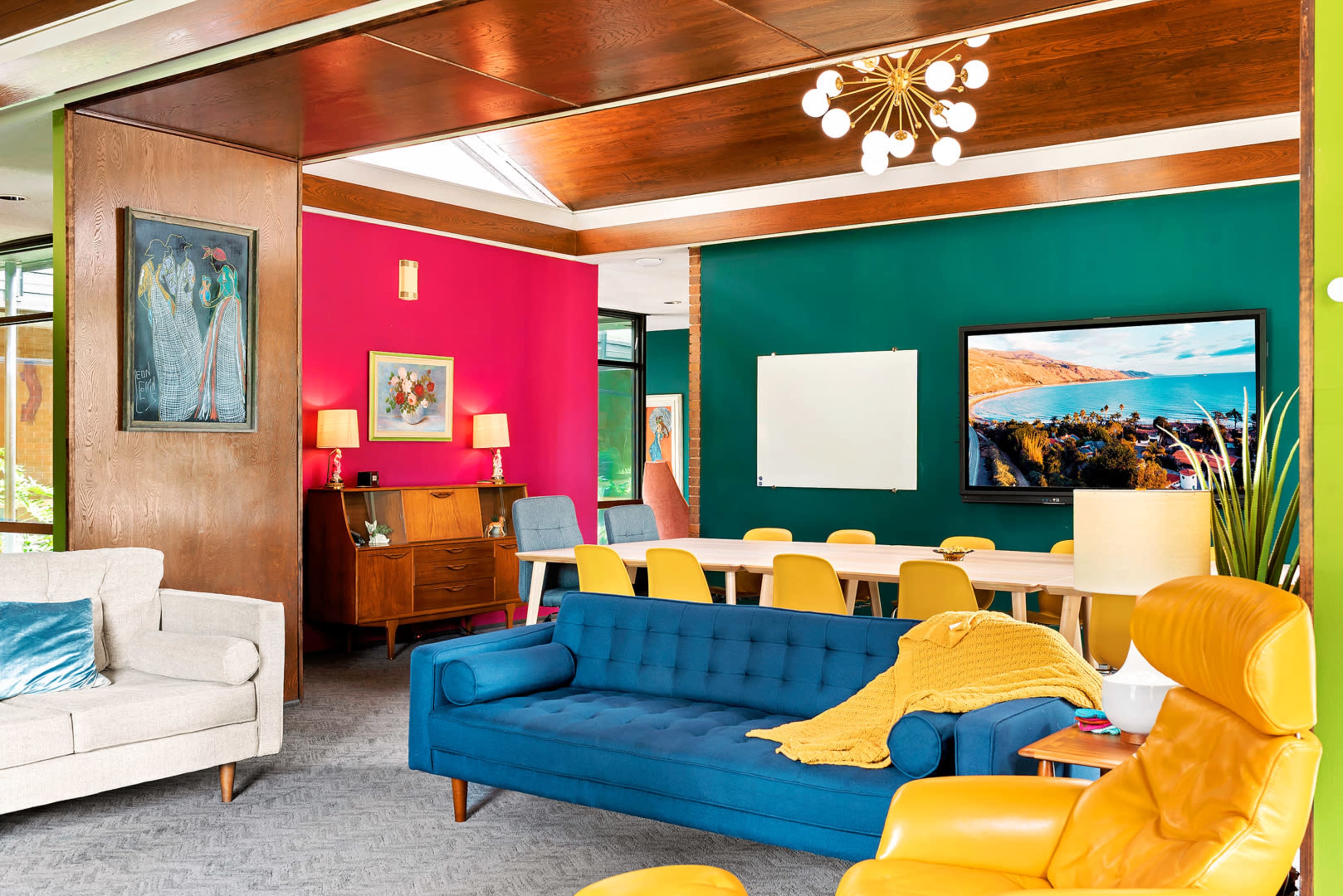 The image depicts a modern interior living space featuring a brightly colored lounge area with a blue sofa, a yellow chair, and walls painted in pink and green, along with a wooden cabinet, framed art, and a large television.