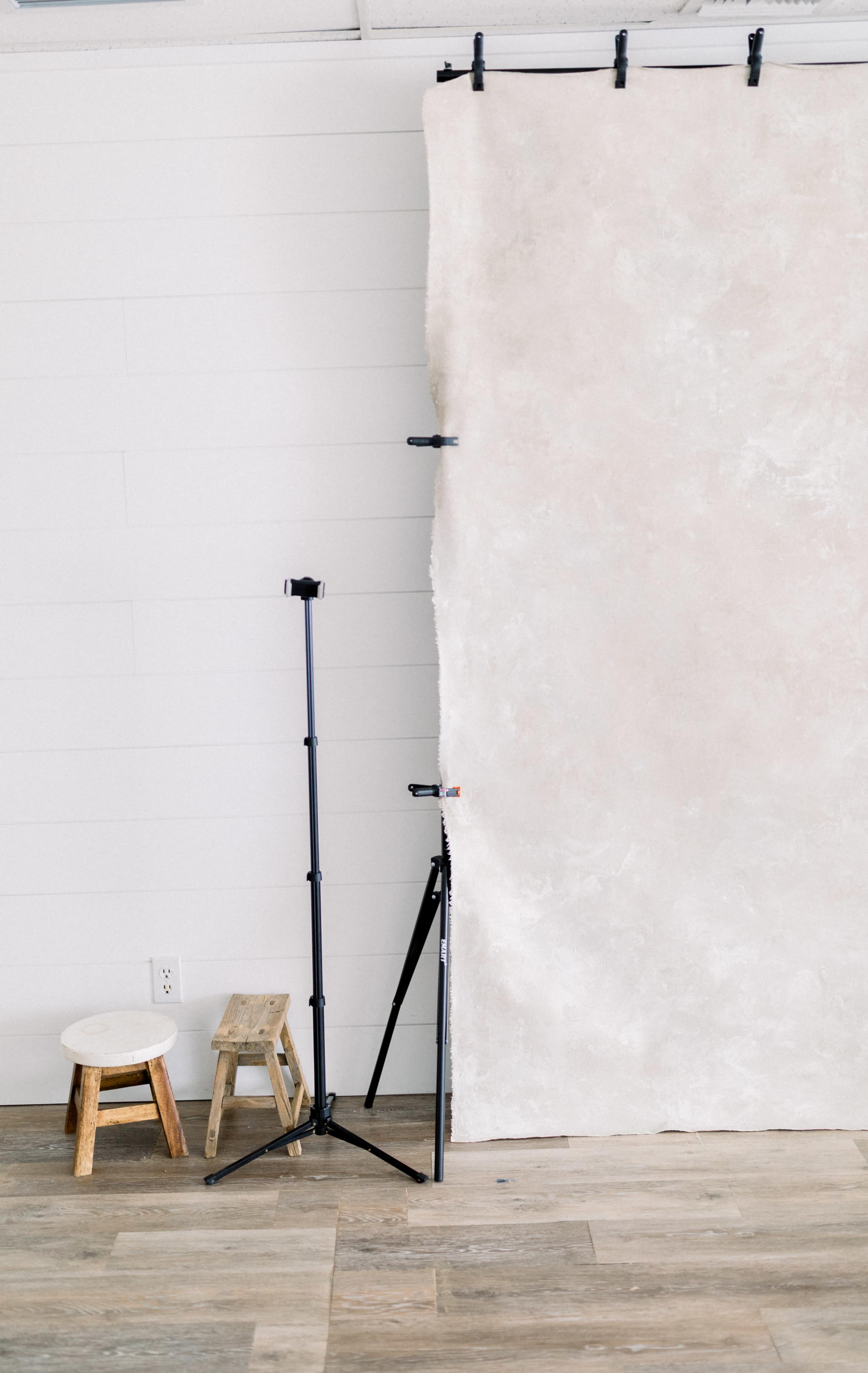 A simple photography backdrop hangs from a black support system, accompanied by a stool and a small wooden step stool on a wooden floor.