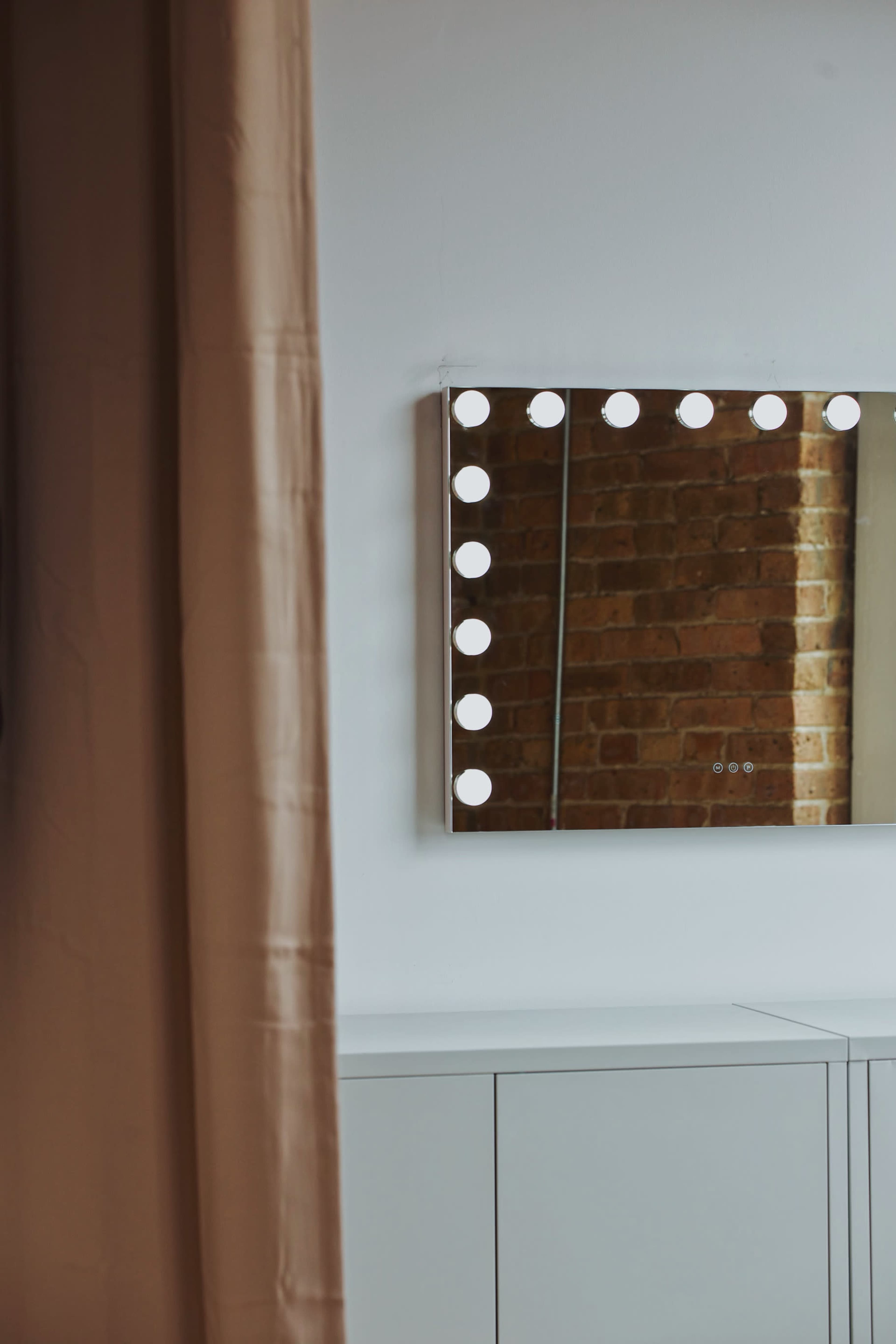 A rectangular mirror with bright circular lights is mounted on a wall next to a beige curtain and a white cabinet.