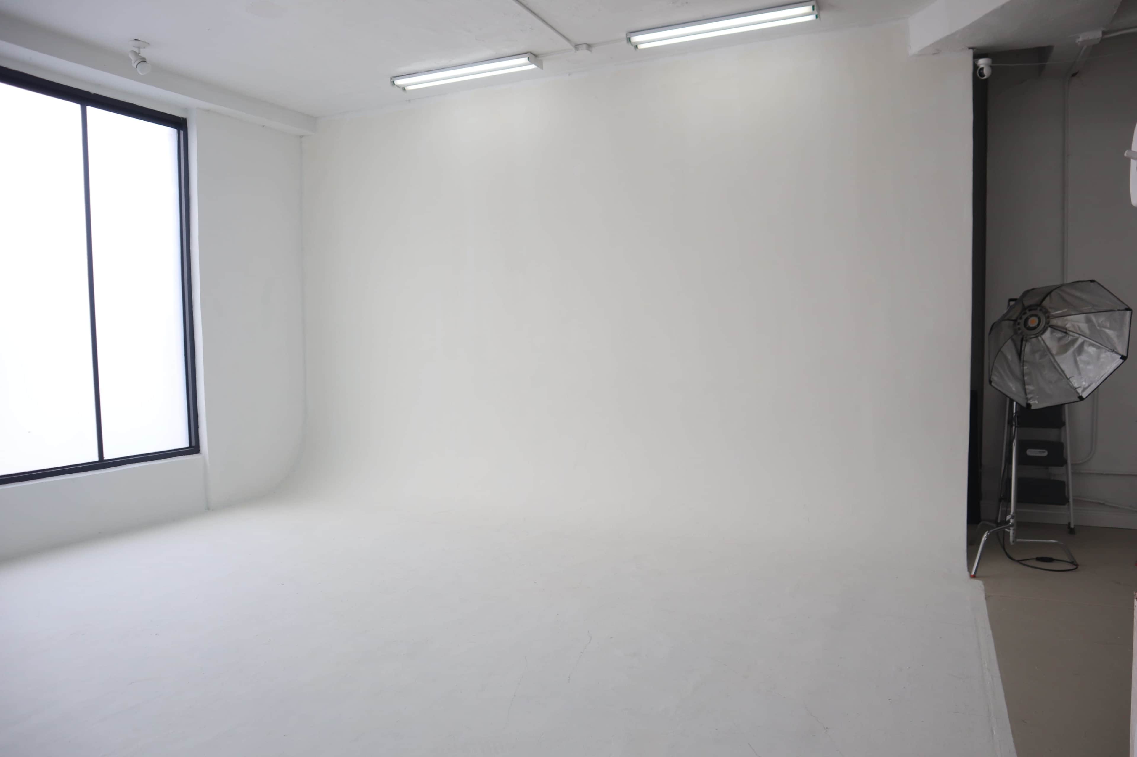 The image shows a minimalist studio space with a white backdrop, large window, and a softbox lighting setup.