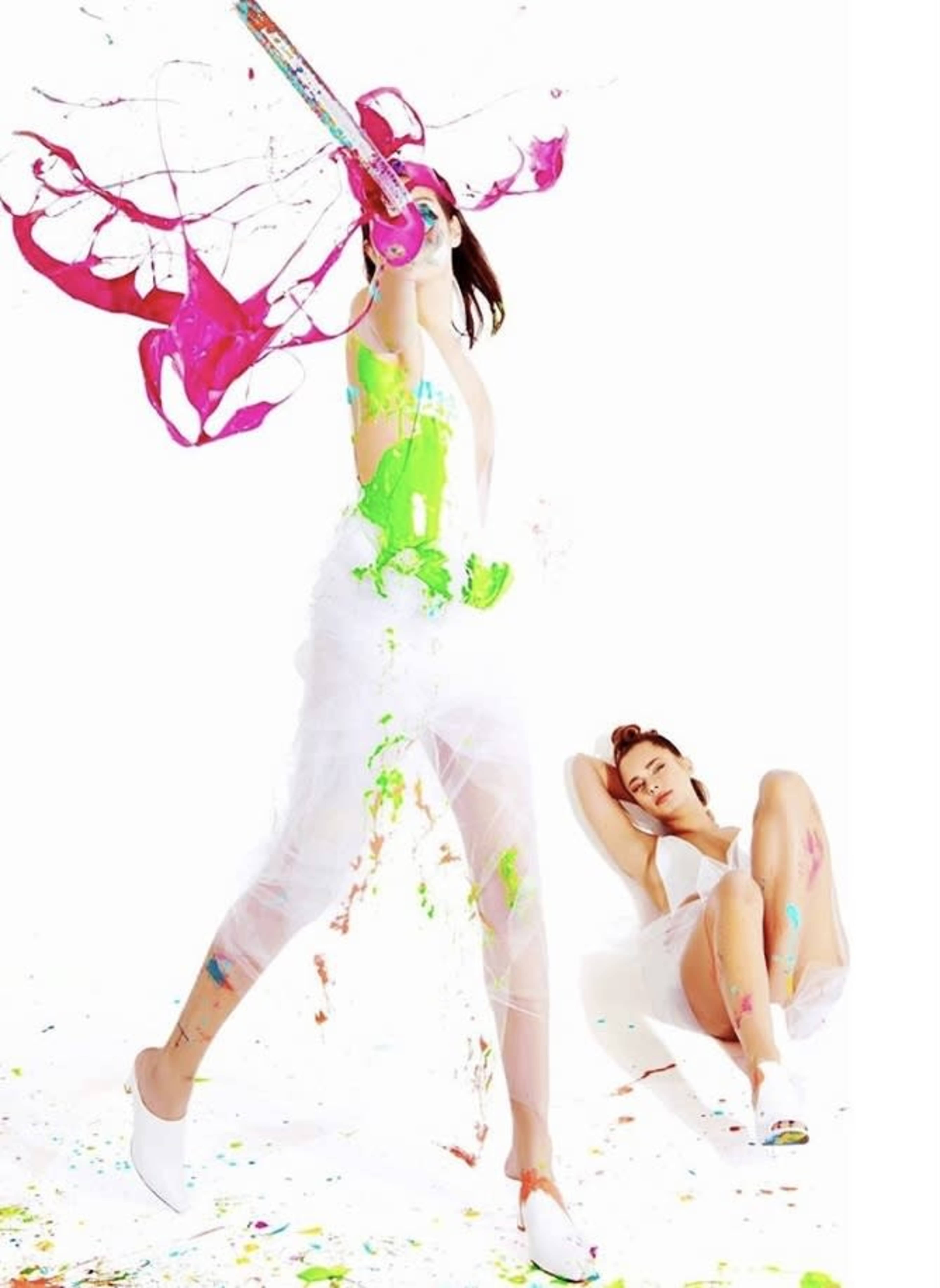 A person dressed in colorful paint is playfully splashing paint while another person sits nearby, appearing relaxed.