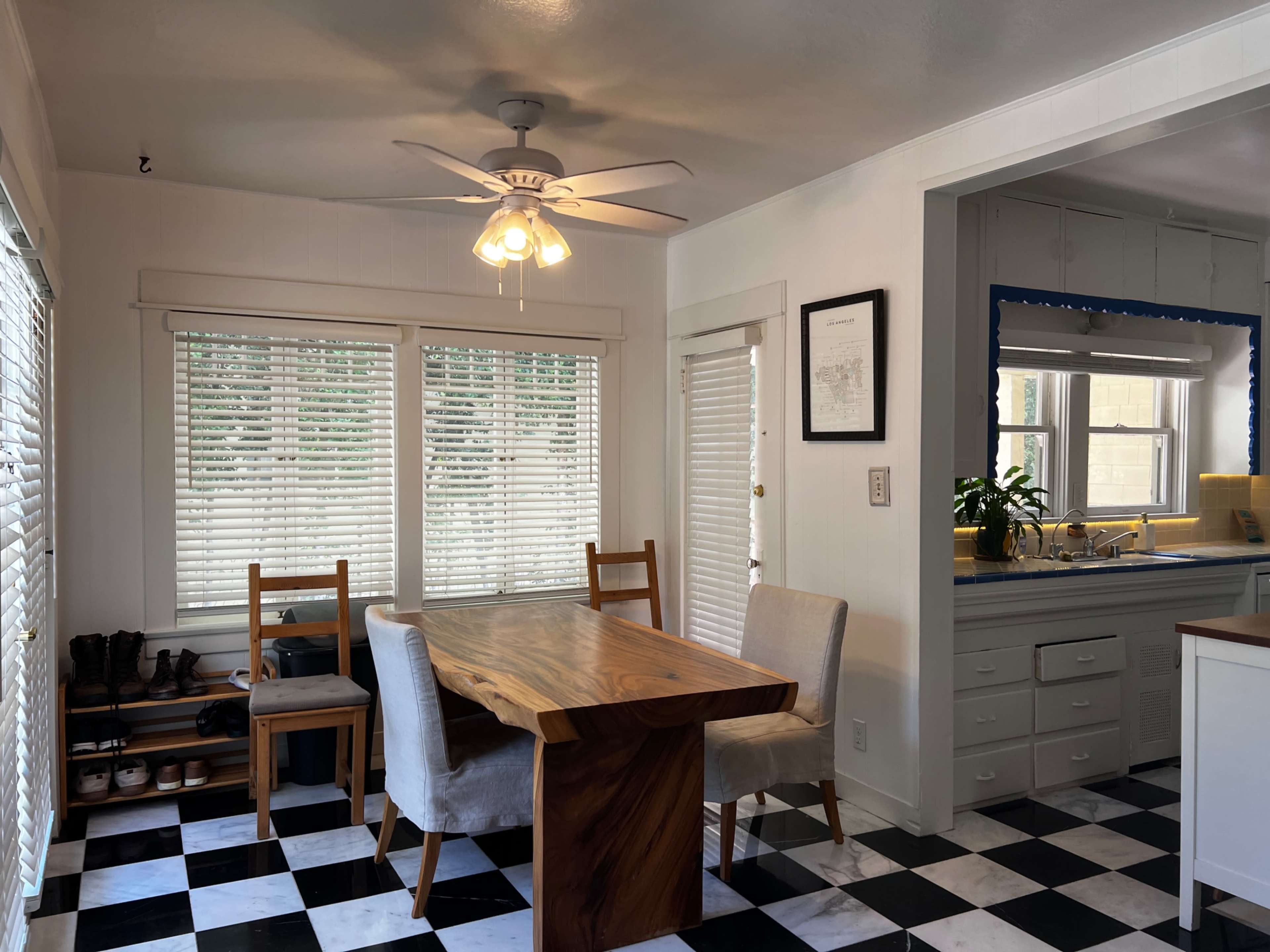 The image shows a dining area with a wooden table and four chairs, surrounded by windows with white blinds, and a kitchen visible in the background with light-colored cabinetry.
