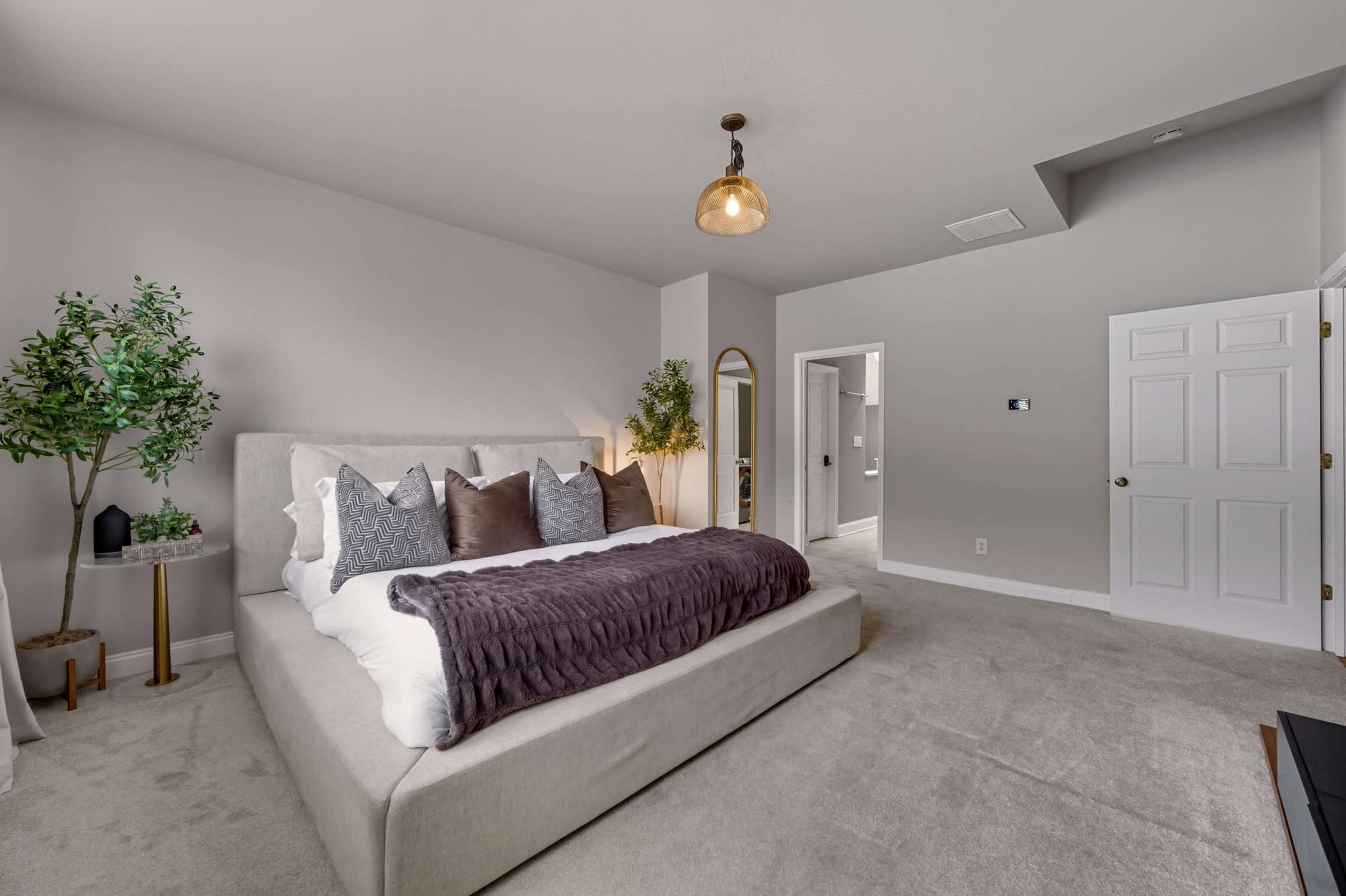 The image shows a spacious, modern bedroom featuring a large bed with decorative pillows, a plant, and an adjoining bathroom visible through a doorway.