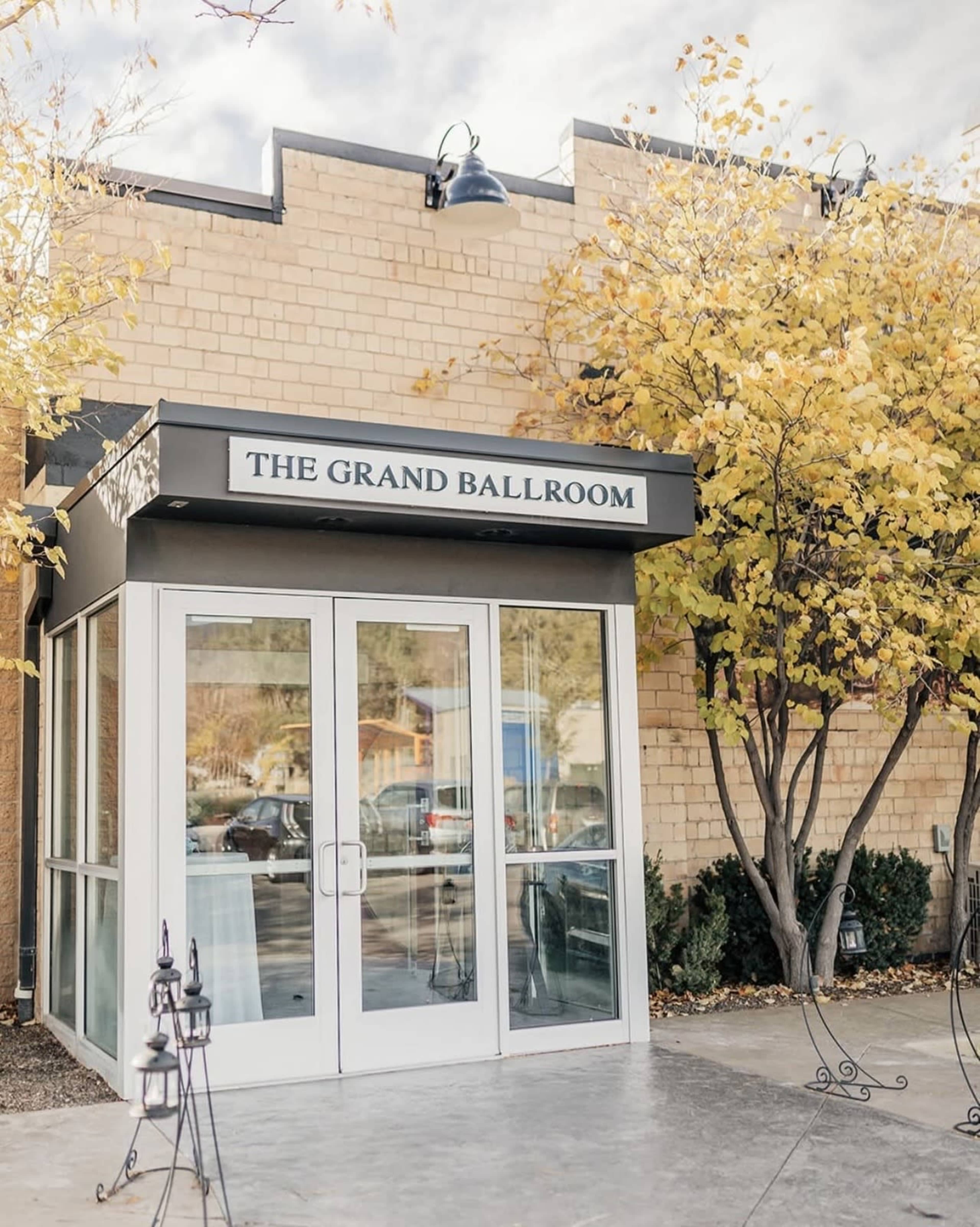 The image shows a modern entrance to a venue called "The Grand Ballroom," framed by yellow-leaved trees and outdoor decor.