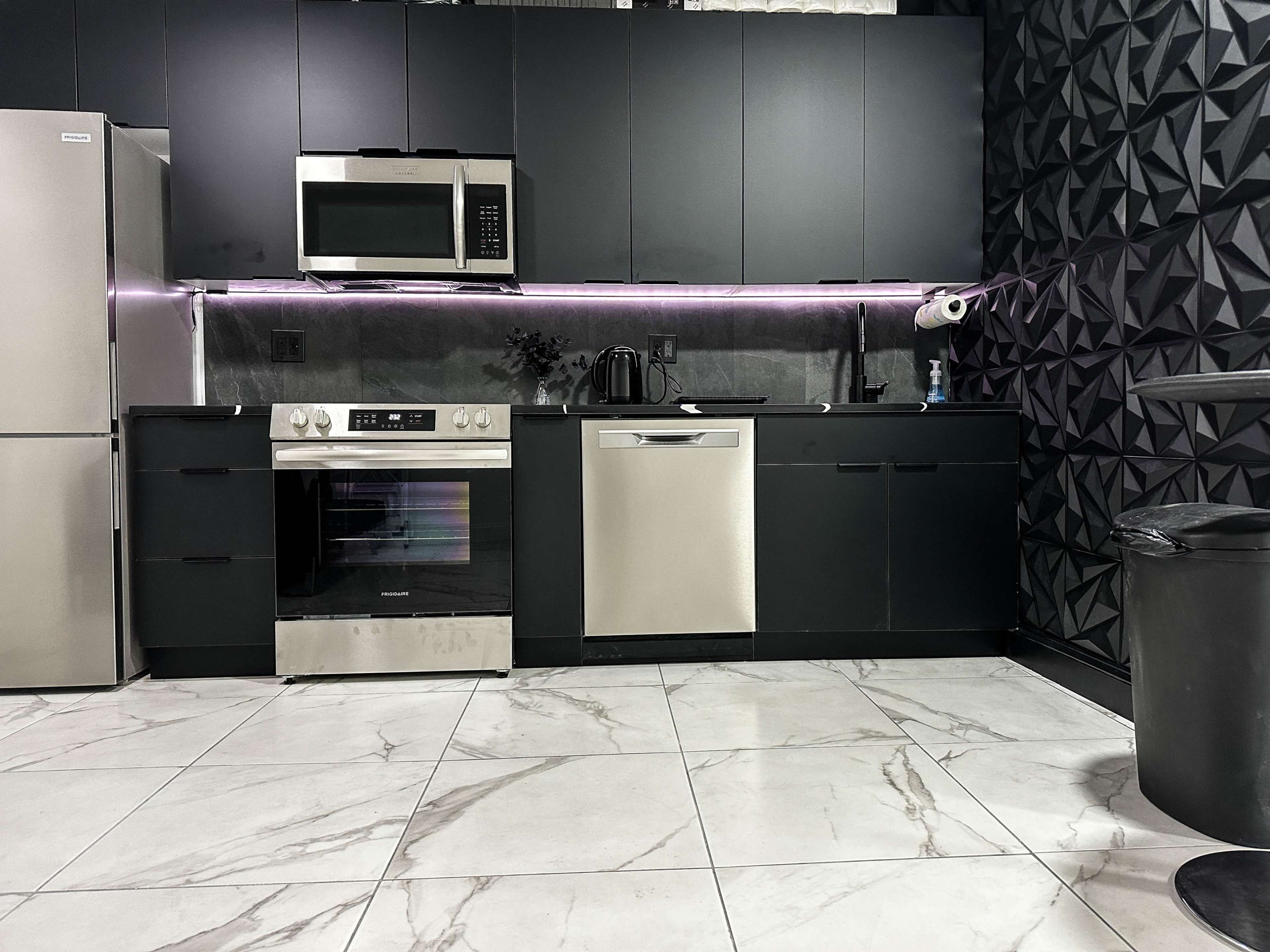 The image shows a modern kitchen featuring black cabinetry, a stainless steel microwave, an oven, and a dishwasher, with a marble floor and geometric wall design.