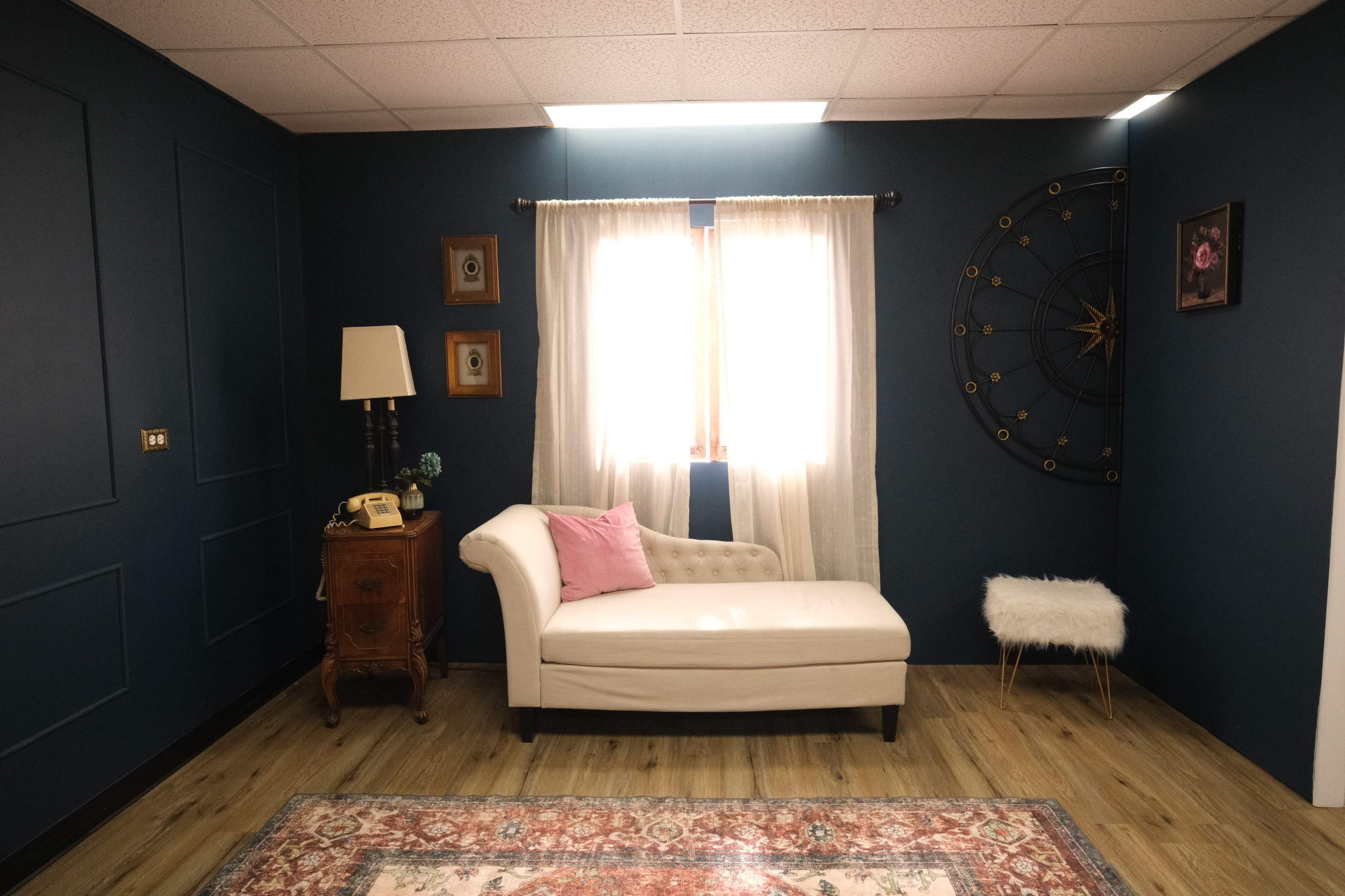 The room features a white chaise lounge, a vintage telephone on a wooden side table, and a large wall clock, all set against dark blue walls and a patterned area rug.