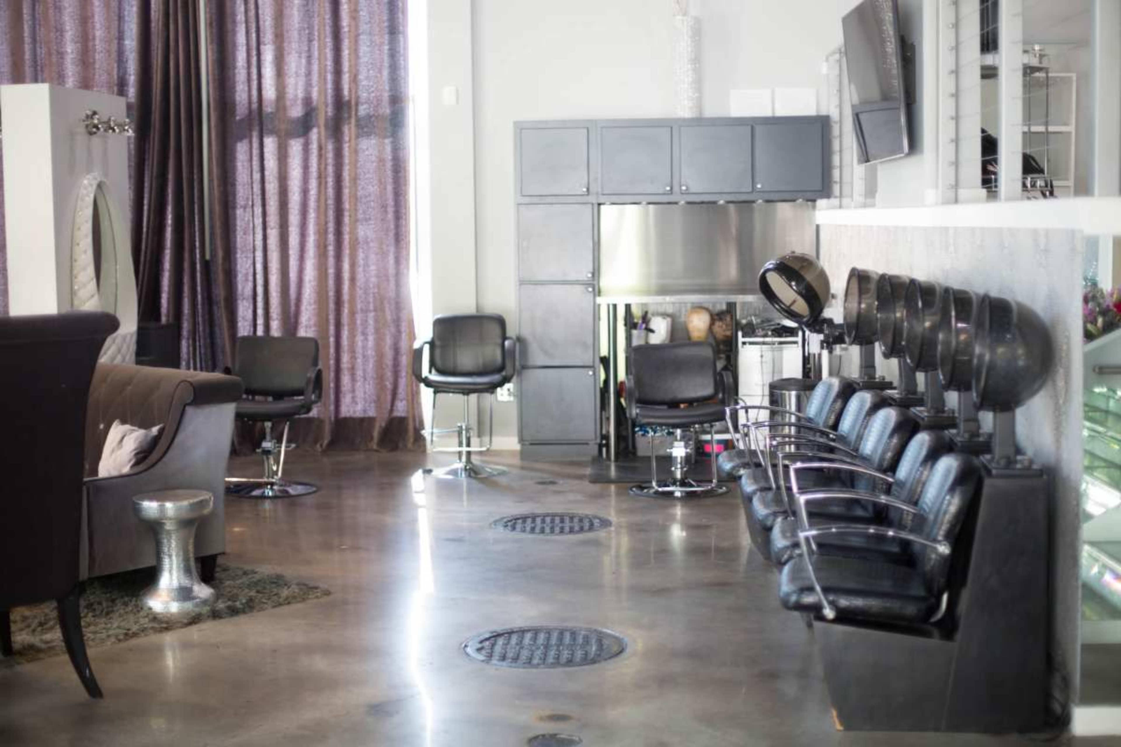 The image shows a modern hair salon with styling chairs, a waiting area, and large windows covered by sheer curtains.