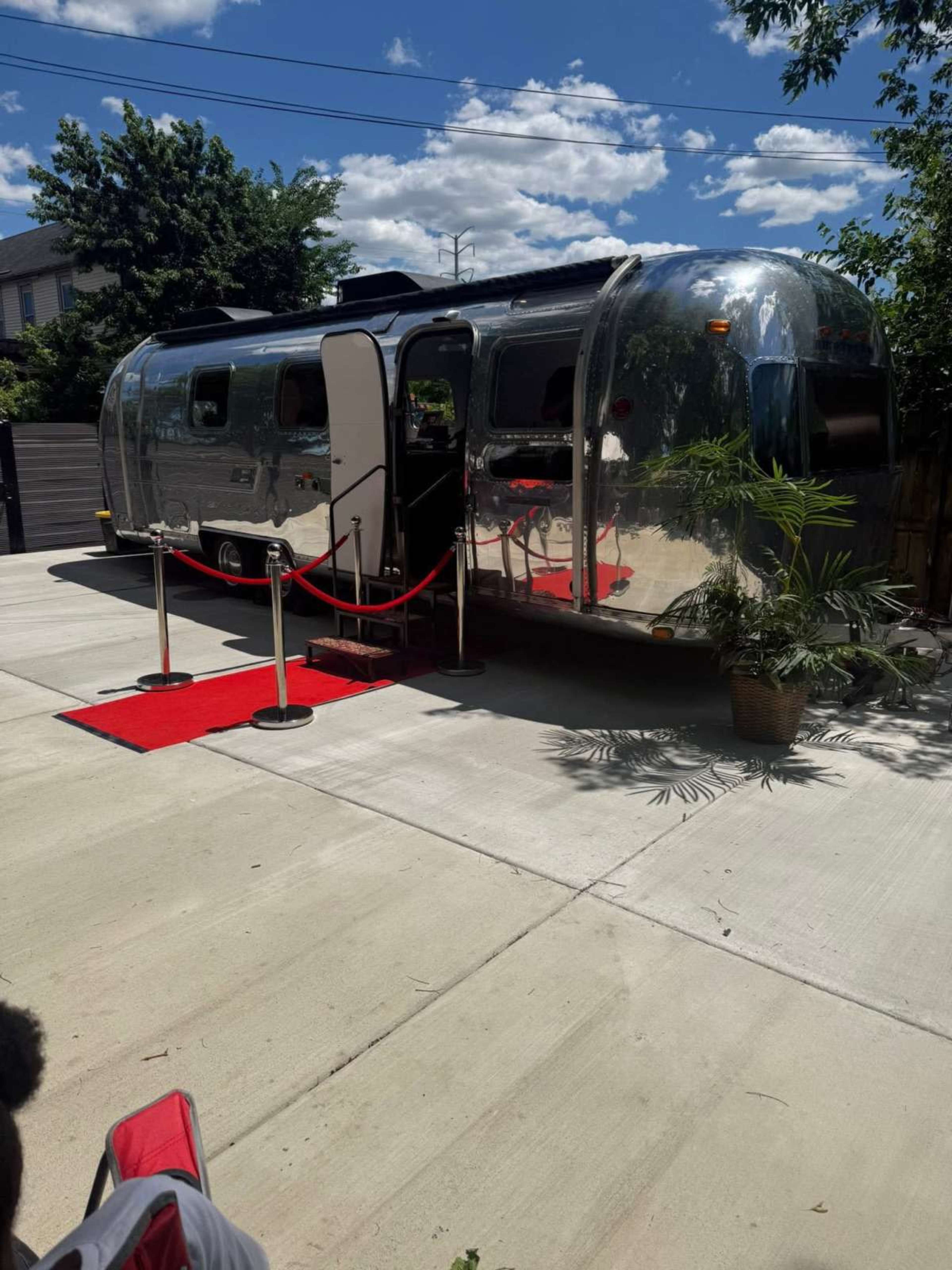 An Airstream trailer is parked on a concrete driveway, featuring red carpet and velvet ropes at its entrance.