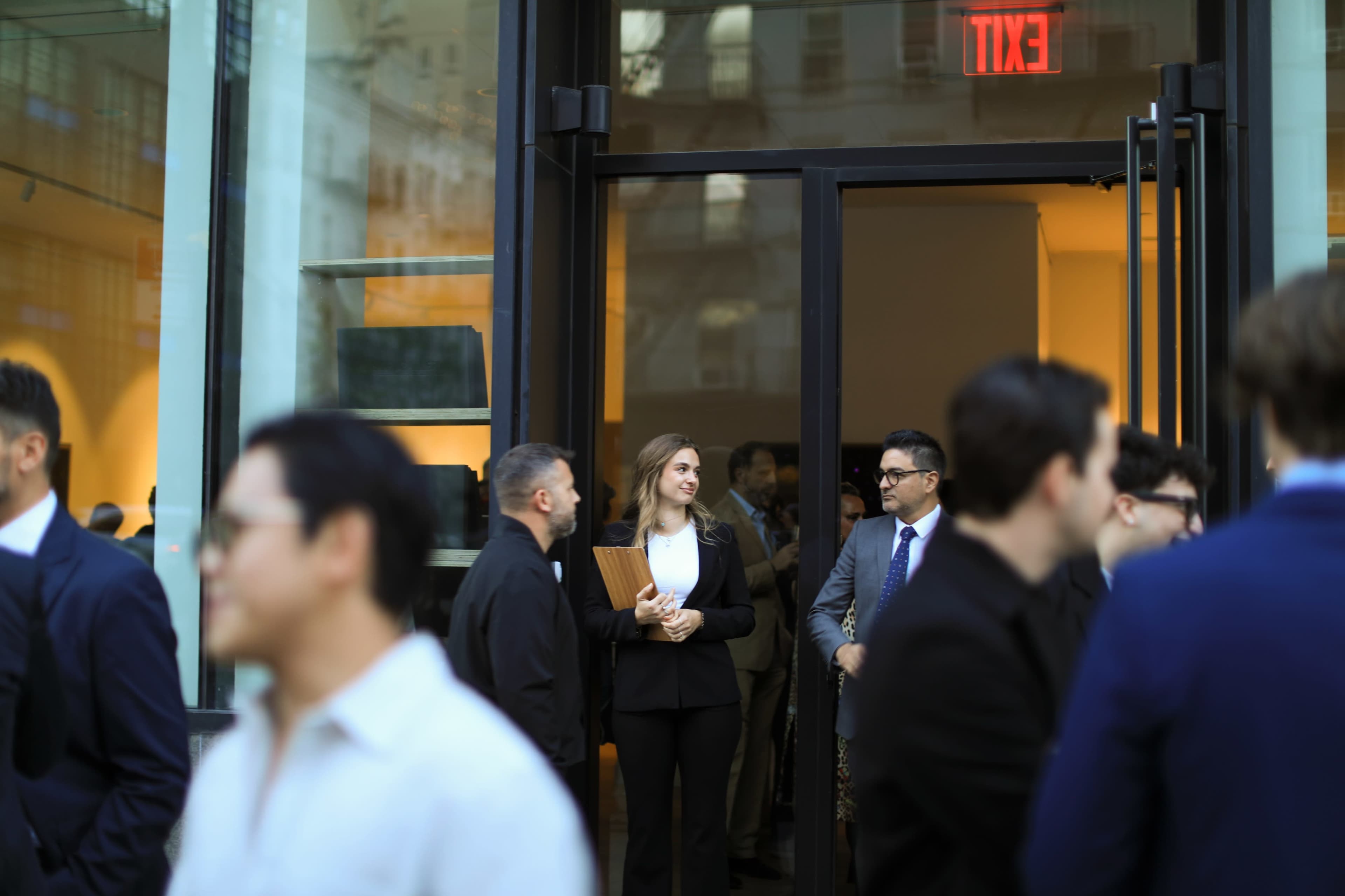 A group of people in business attire mingles outside a glass entrance, while a woman stands at the door holding a clipboard.