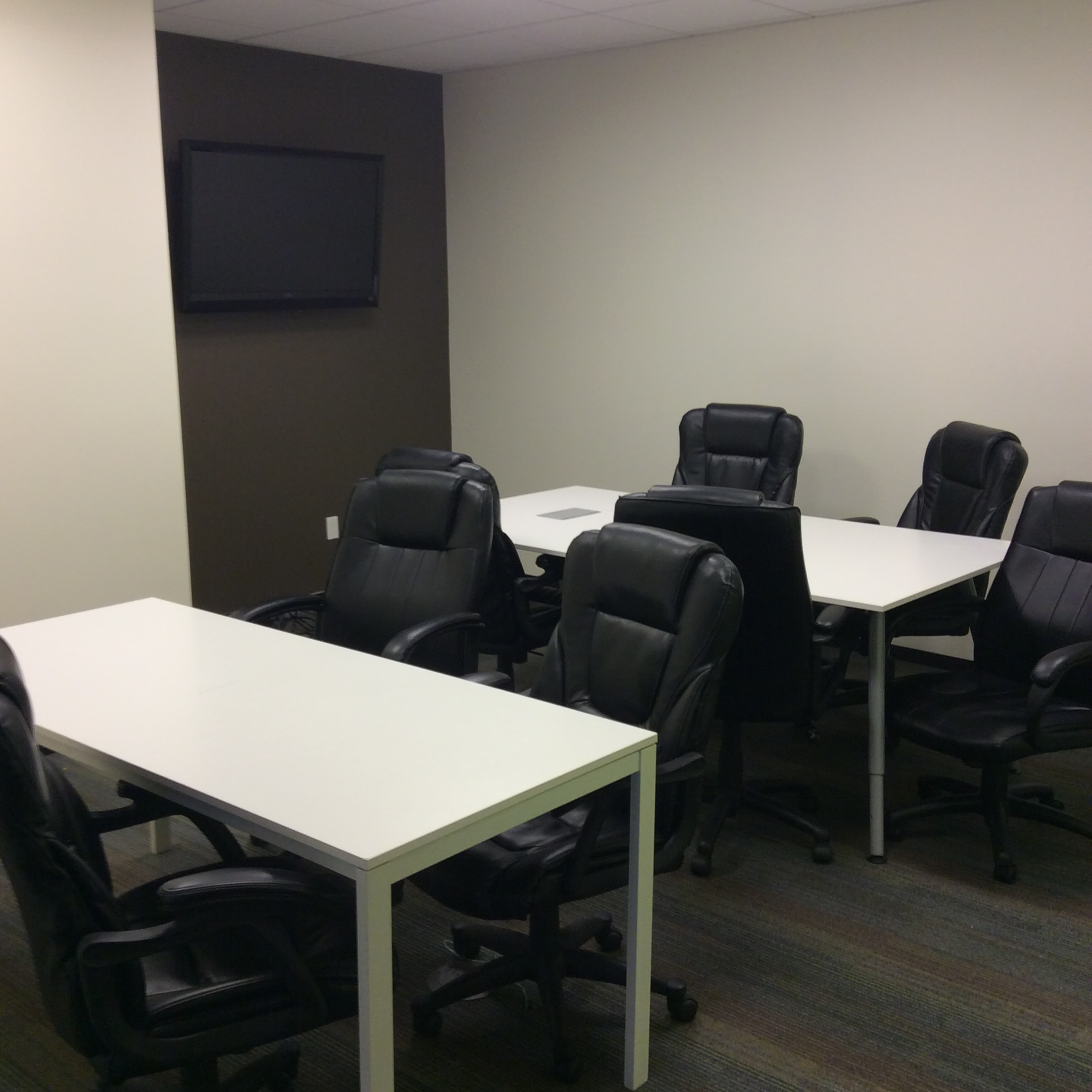 A conference room with two white tables surrounded by black office chairs and a mounted television on the wall.