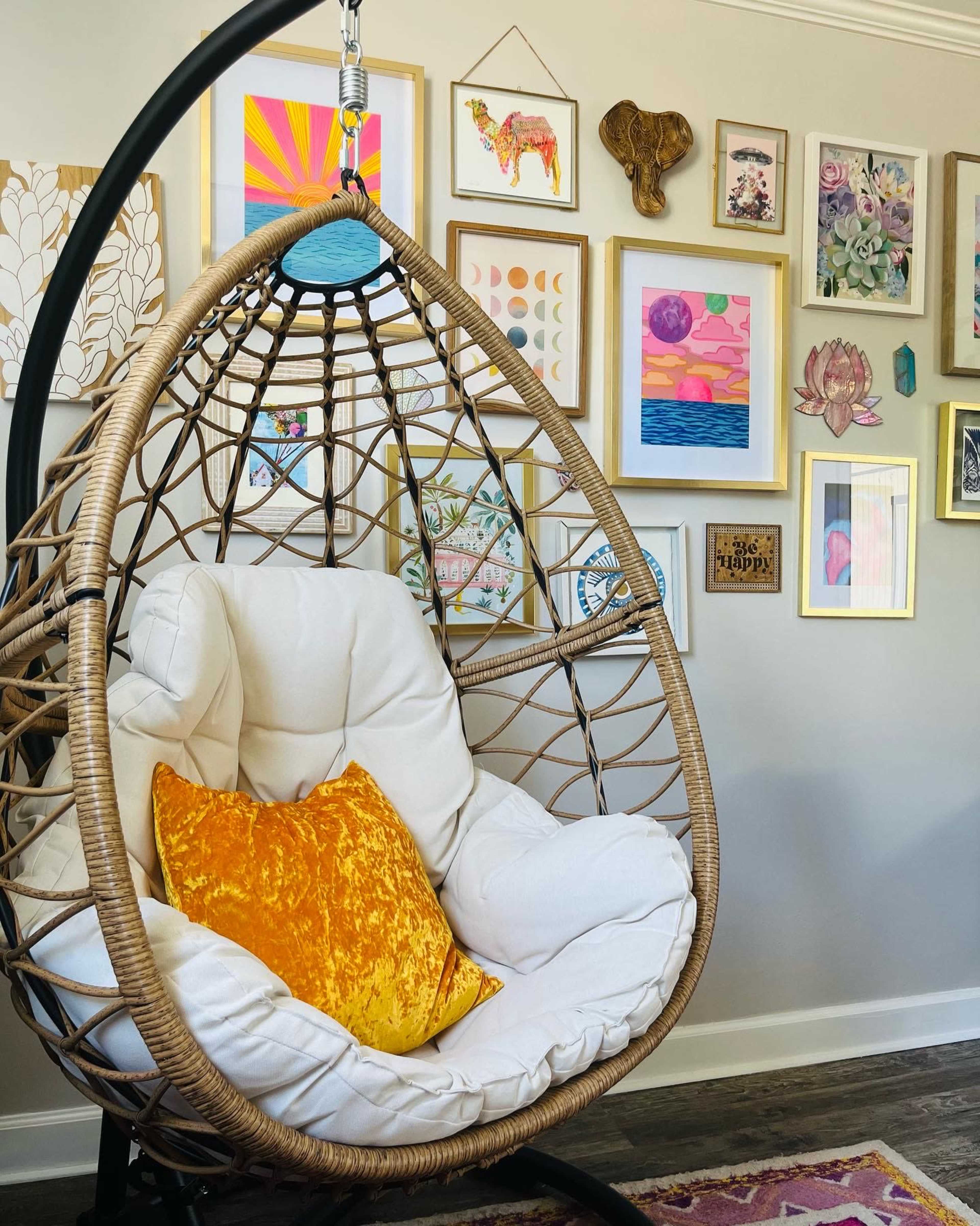 A hanging rattan chair with a white cushion and an orange pillow, surrounded by an arrangement of framed artwork on the wall.