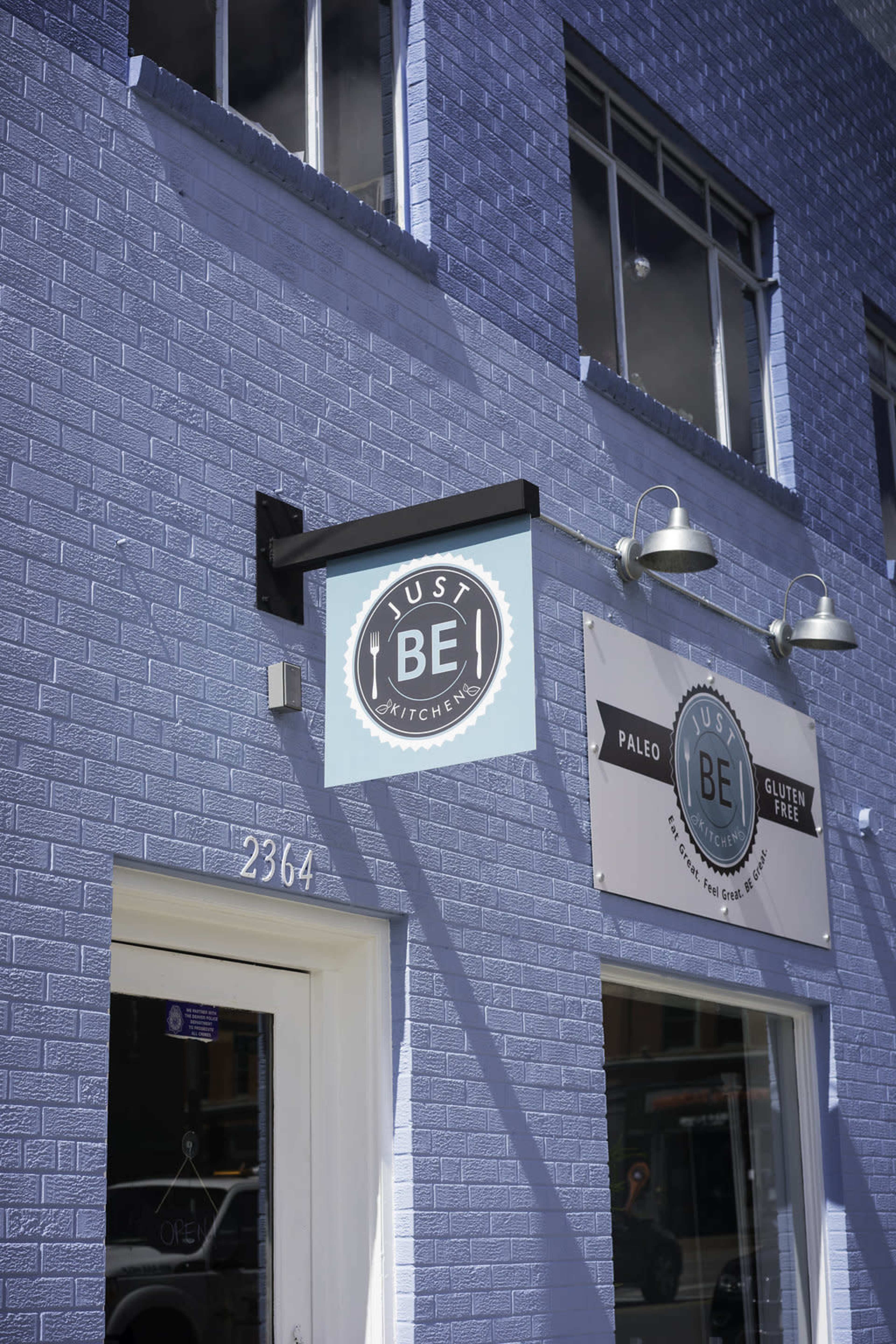 The exterior of a restaurant named "Just Be Kitchen," featuring a light blue brick wall and two signs indicating it offers paleo and gluten-free options.