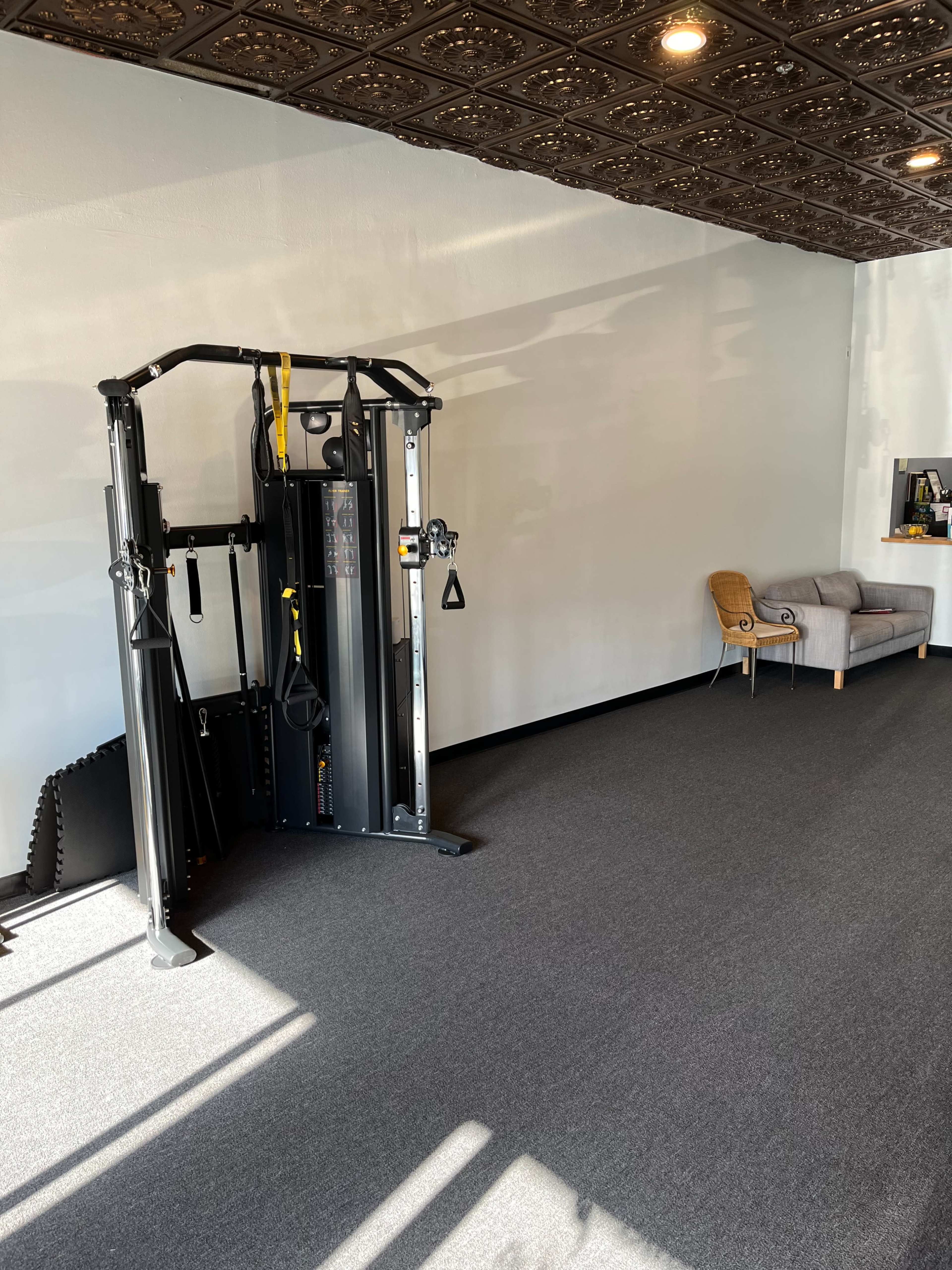 The image shows a gym interior featuring a cable machine on the left and a gray chair positioned against a wall on the right.