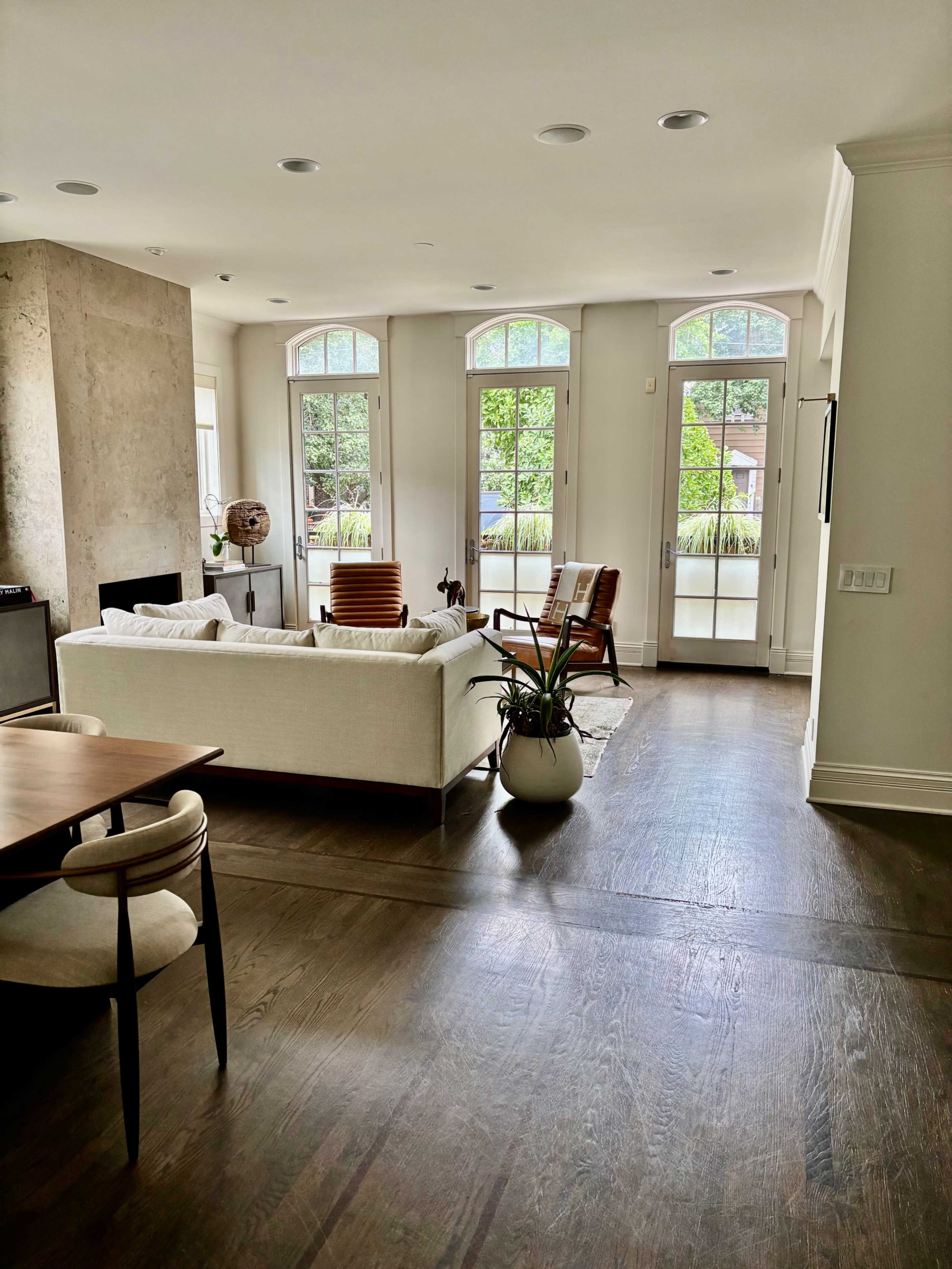 The image shows a modern living room with large windows, a white sofa, a wooden dining table, and a potted plant.