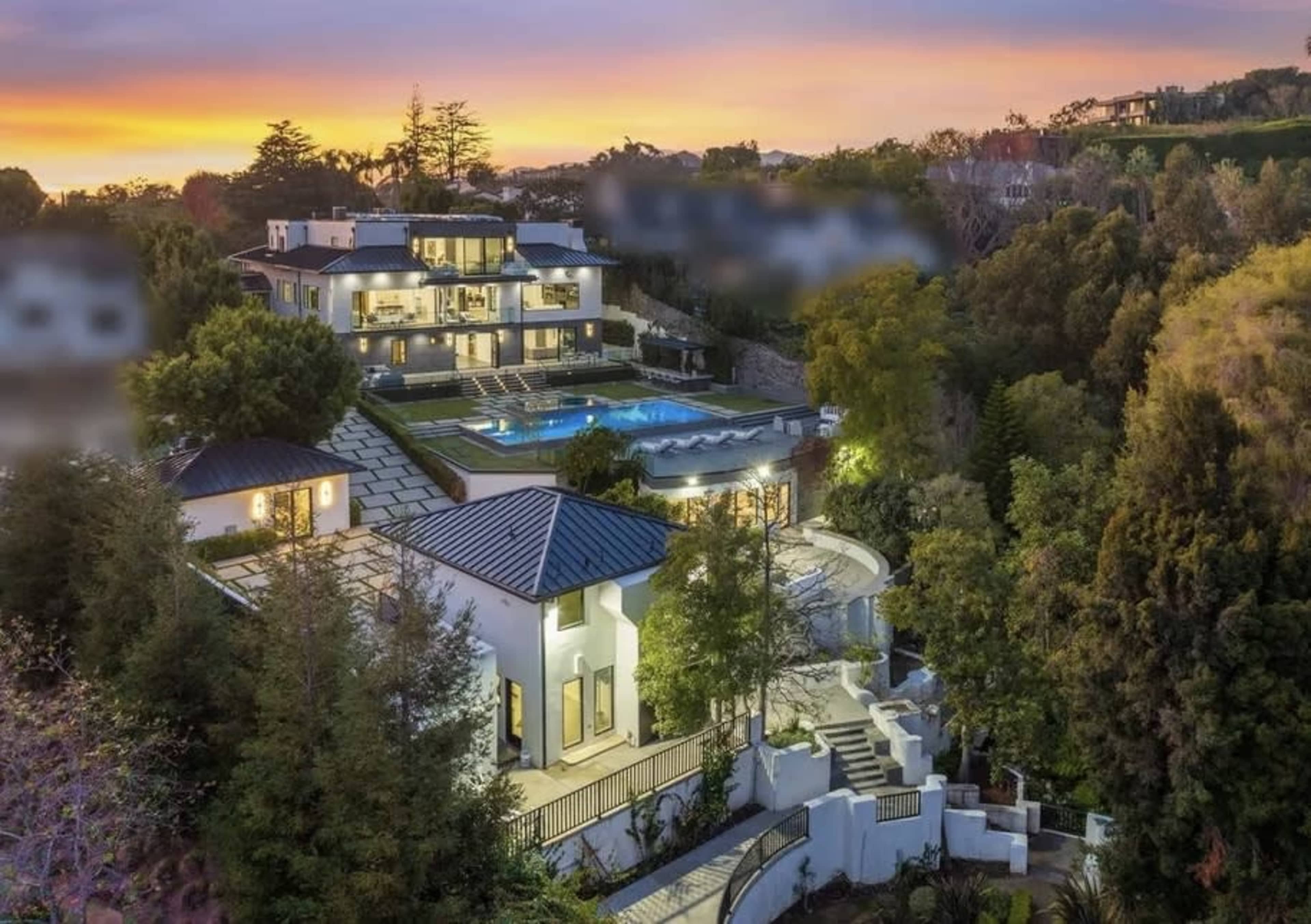 A large, modern hillside mansion with multiple levels, a swimming pool, and extensive landscaping at sunset.