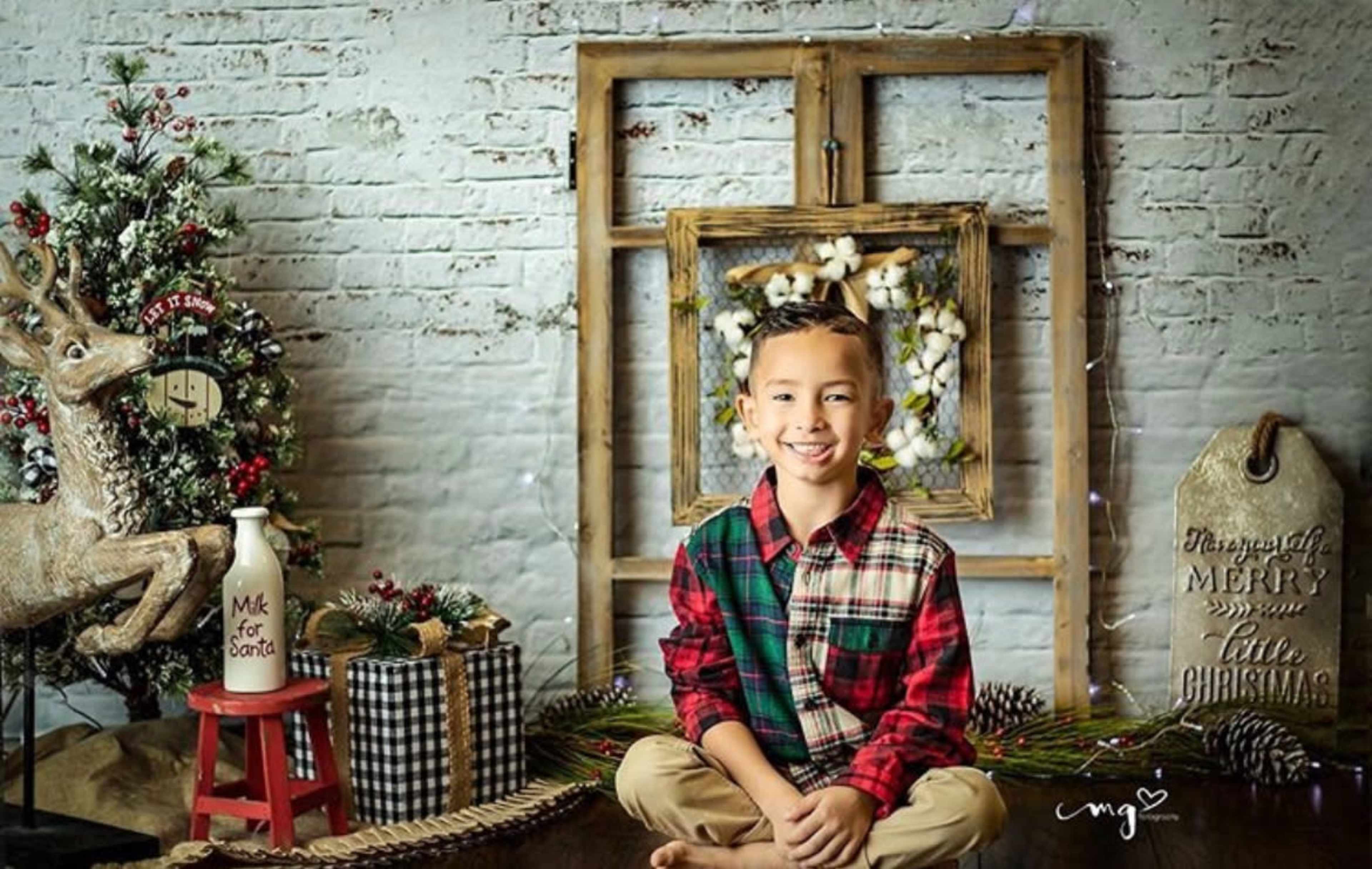 A young boy sits in front of a decorated Christmas scene featuring a tree, a reindeer, and various festive decorations.
